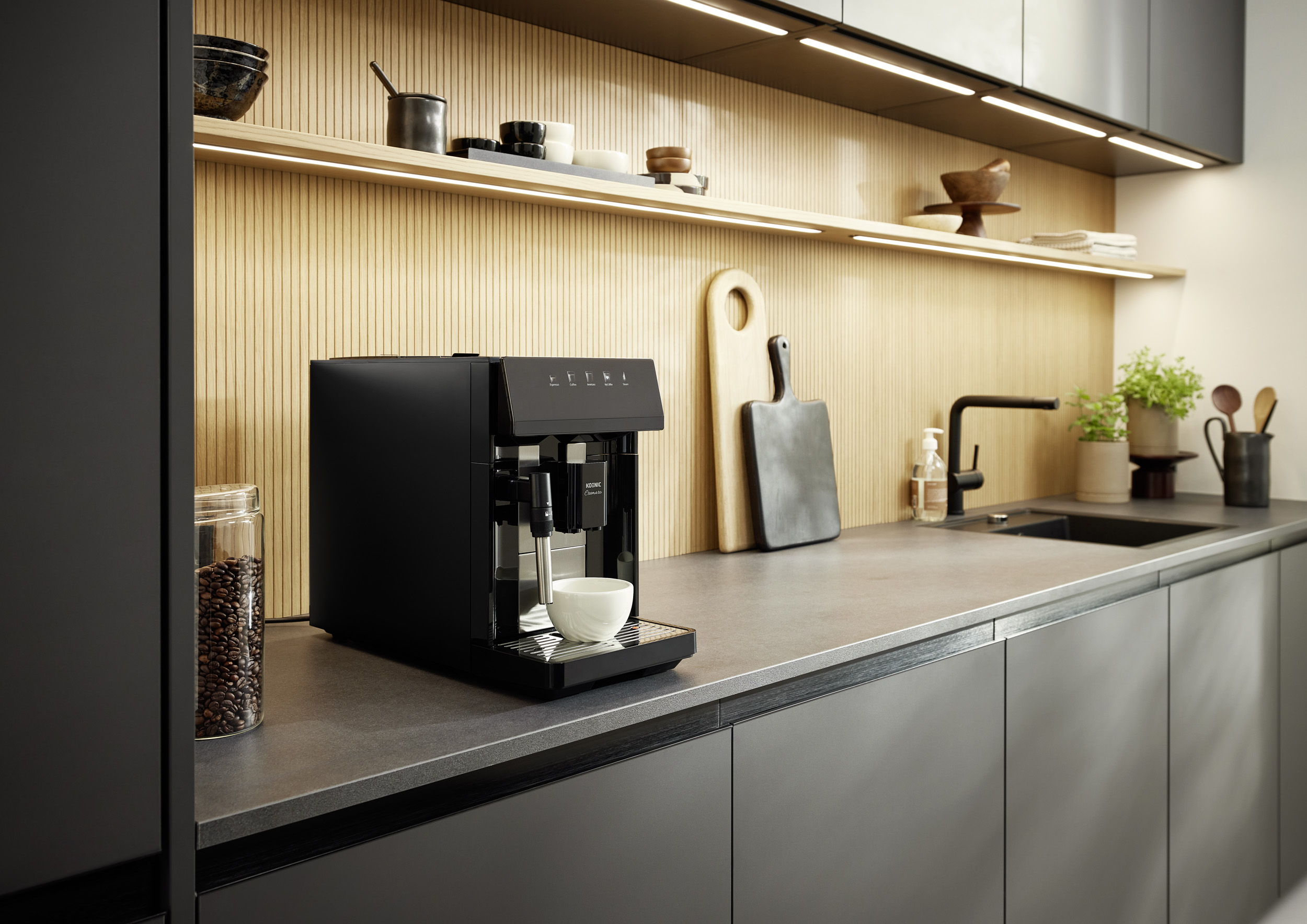 Moderne Küche mit schwarzer Kaffeemaschine auf dunkler Arbeitsplatte, Holzwand und beleuchteten Regalen.