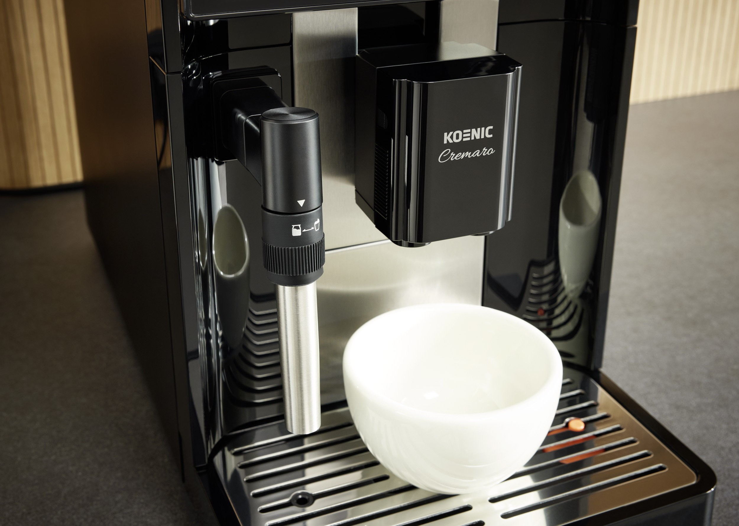 Schwarze Koenic Cremaro Kaffeemaschine mit Milchschäumer und weißer Tasse auf Edelstahl-Abtropfgitter.