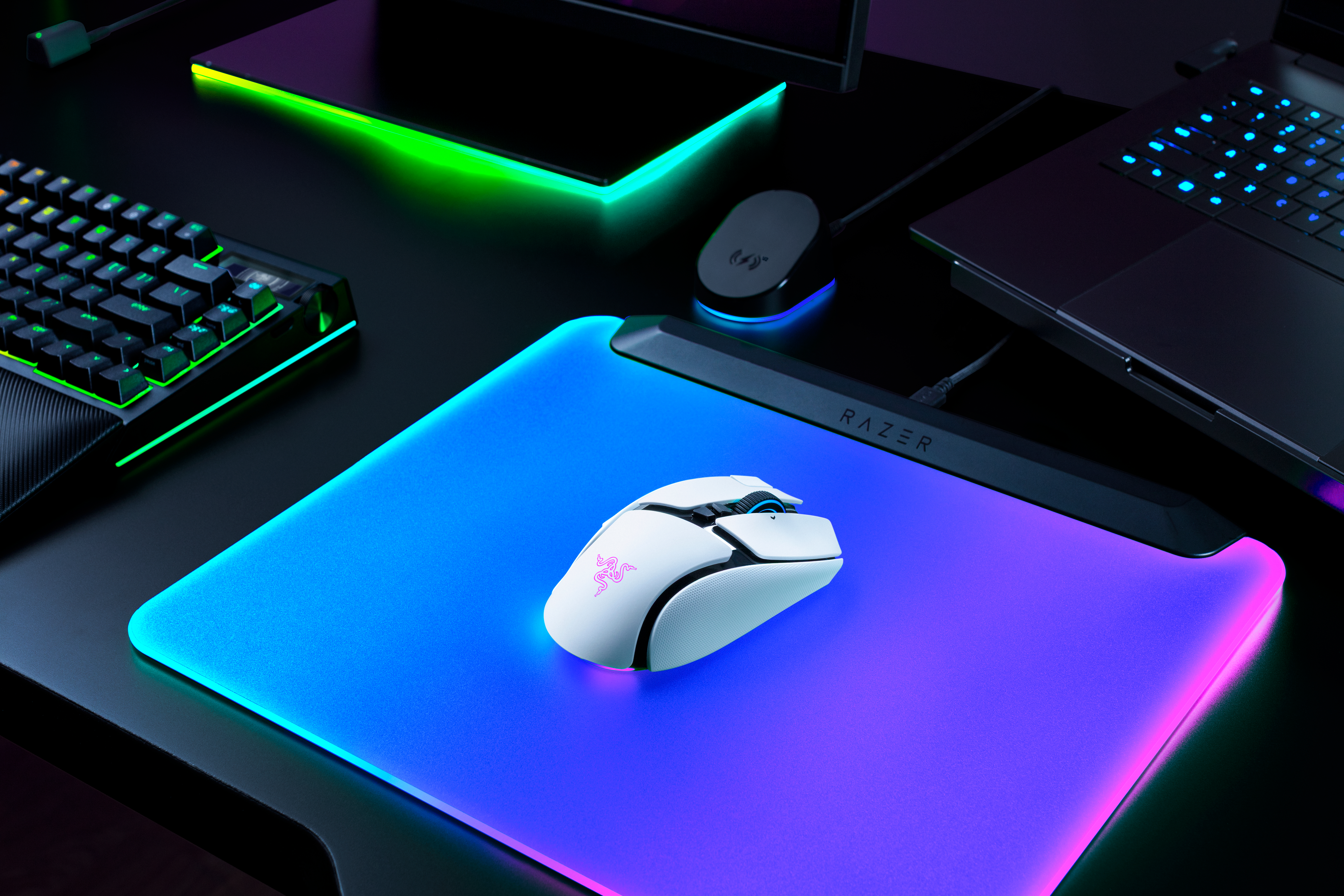 Eine weiße Gaming-Maus mit RGB-Beleuchtung auf einem leuchtenden Razer-Mauspad, umgeben von Tastatur und Laptop.