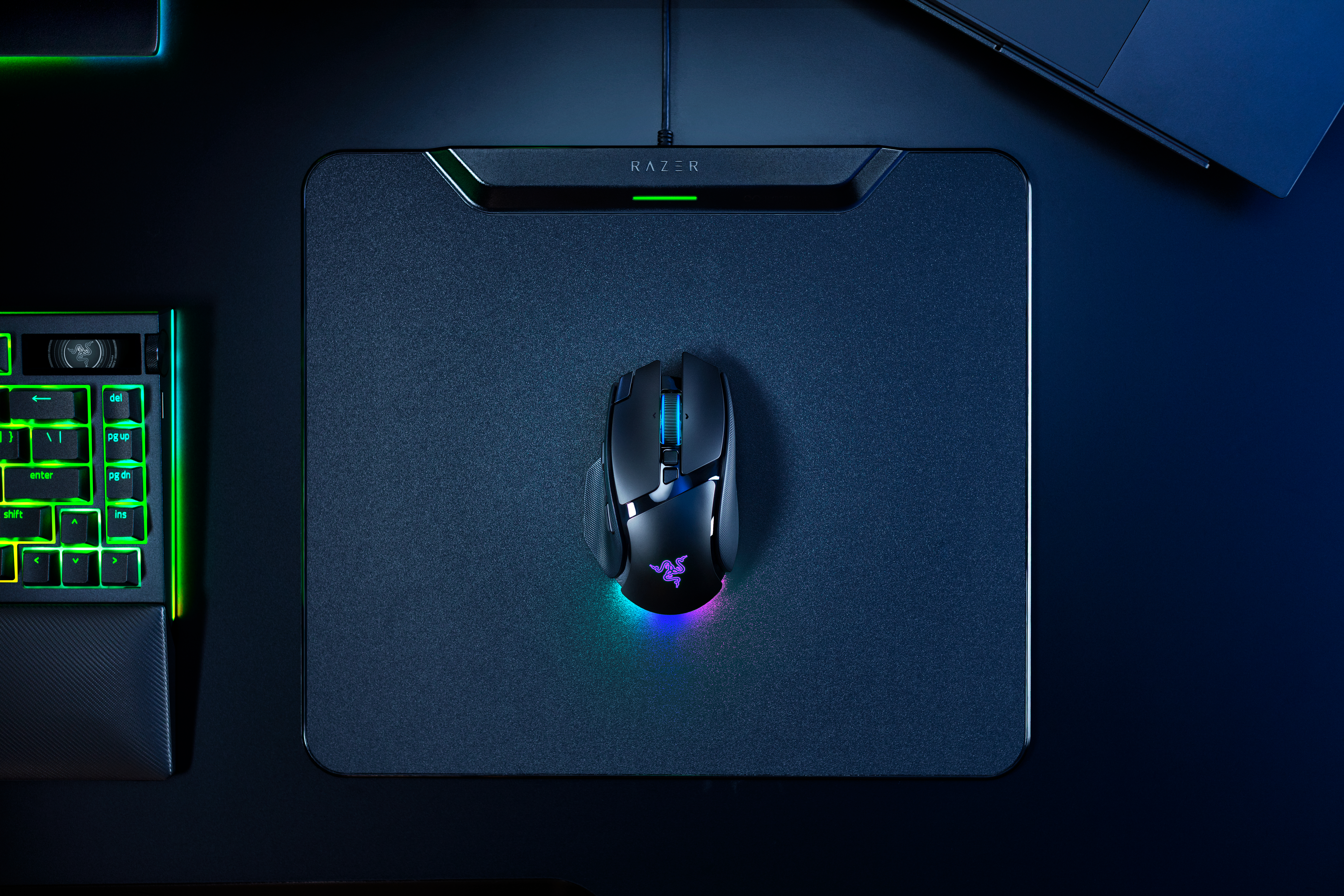 Eine schwarze Razer-Gaming-Maus mit RGB-Beleuchtung auf einem Mauspad, mit Tastatur und Laptop im Hintergrund.