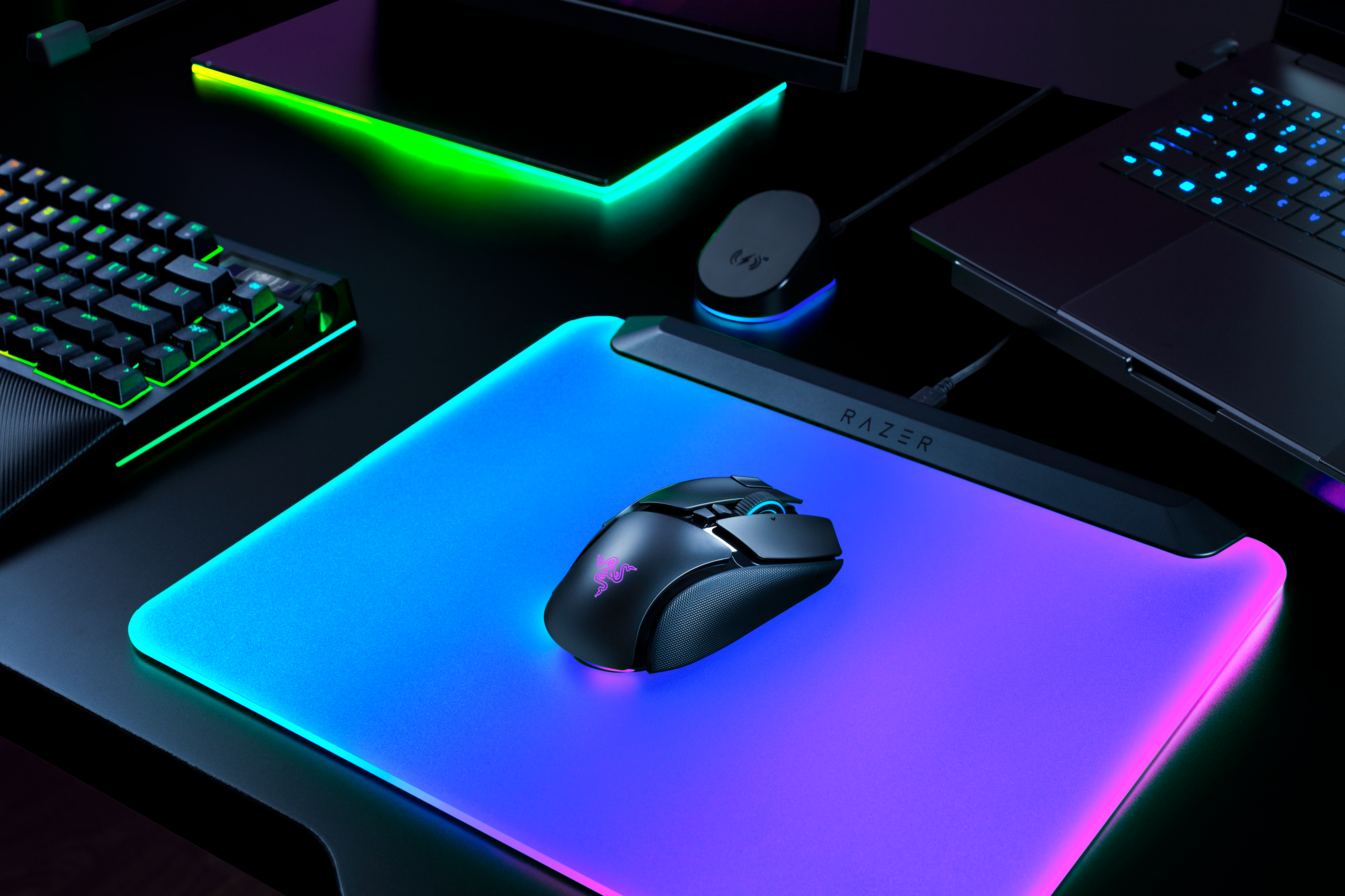 Eine schwarze Razer-Maus mit LED-Beleuchtung auf einem leuchtenden Mauspad, umgeben von Tastatur und Laptop.