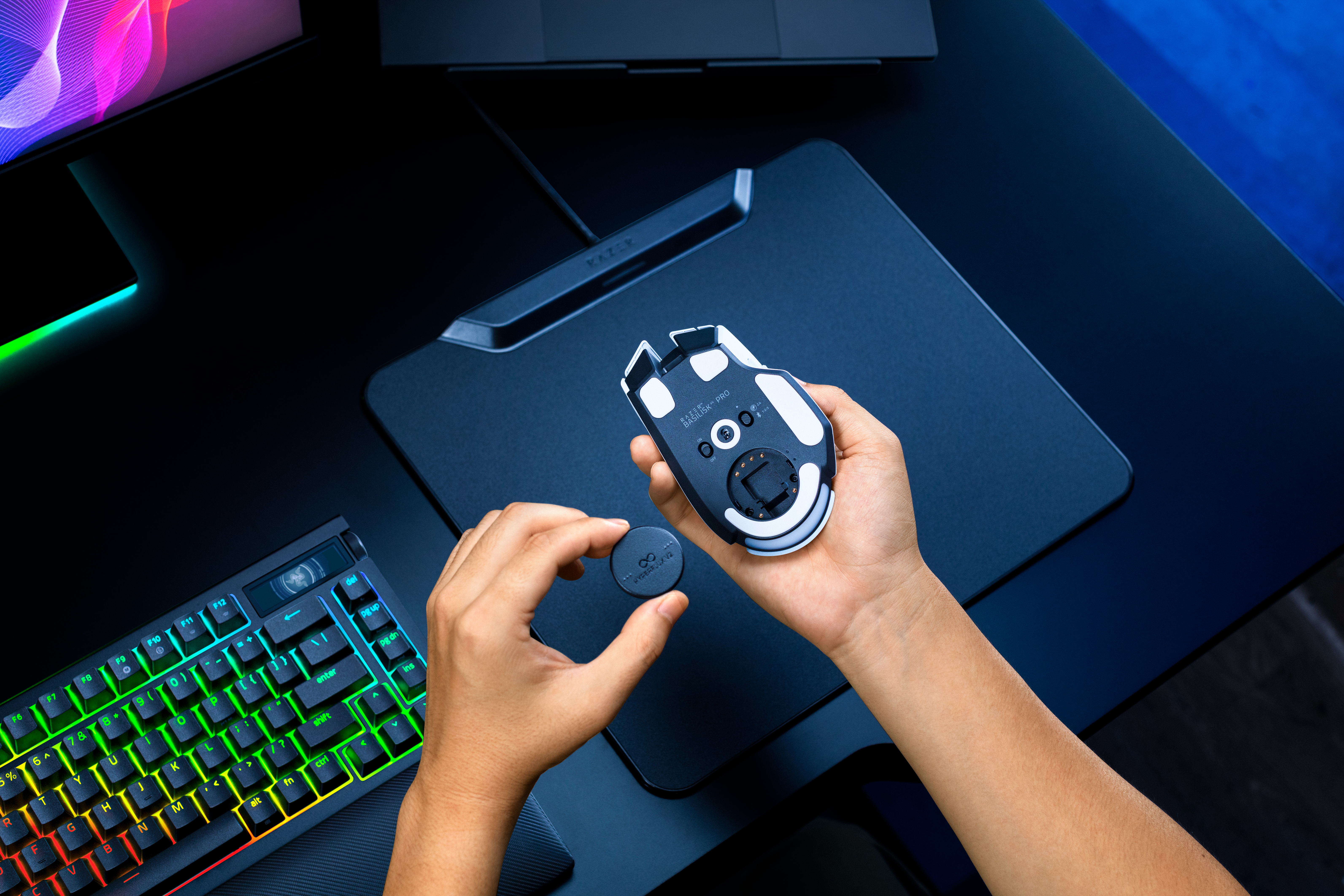 Eine Person hält eine weiße Gaming-Maus und einen runden Chip über einem Schreibtisch mit Tastatur und Mauspad.