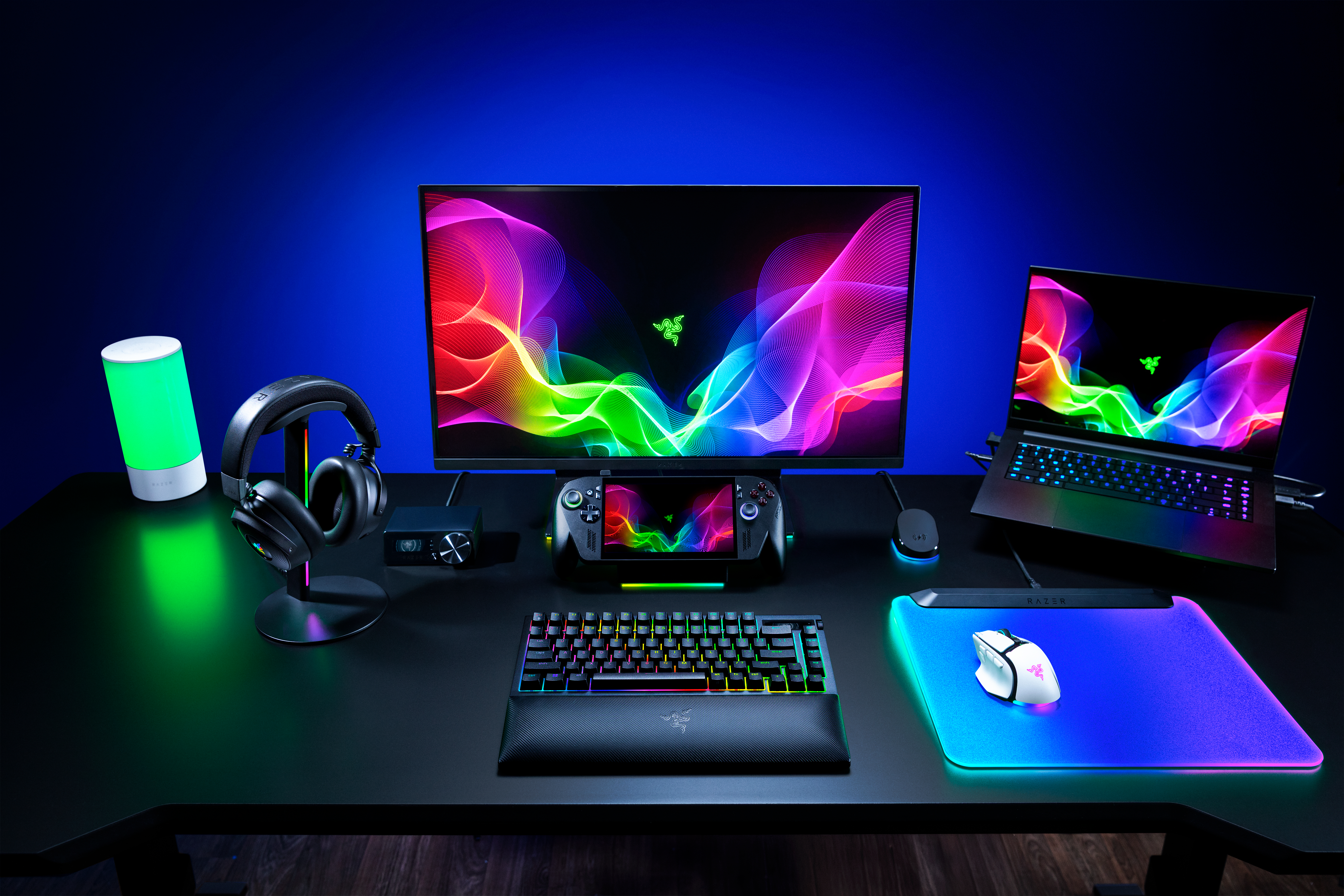 Ein Gaming-Setup auf einem schwarzen Tisch mit bunten Bildschirmen, Tastatur, Maus, Laptop und Kopfhörern.