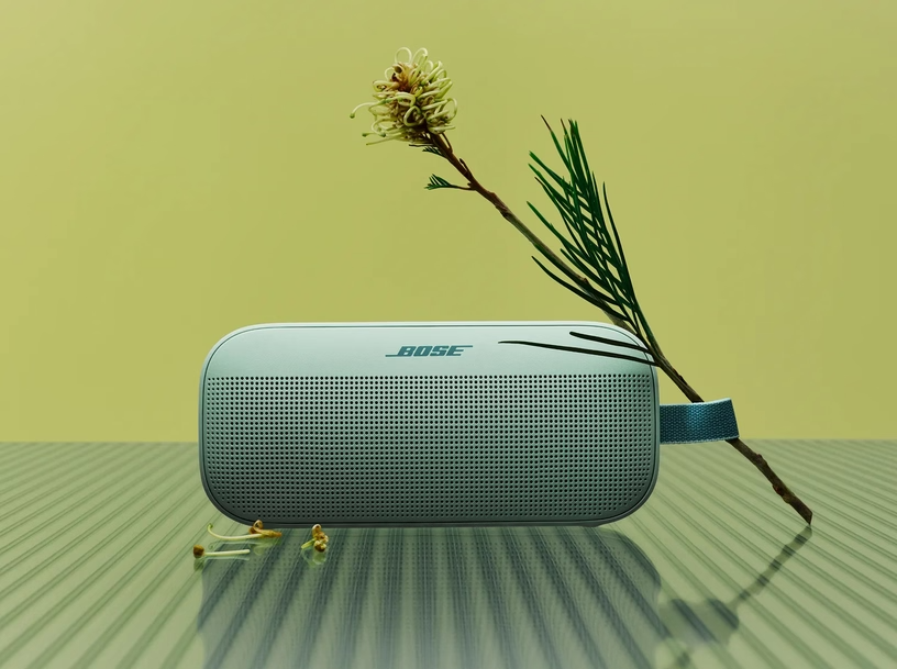Ein blauer Bose-Bluetooth-Lautsprecher steht auf einer gläsernen Oberfläche neben einer Blüte.