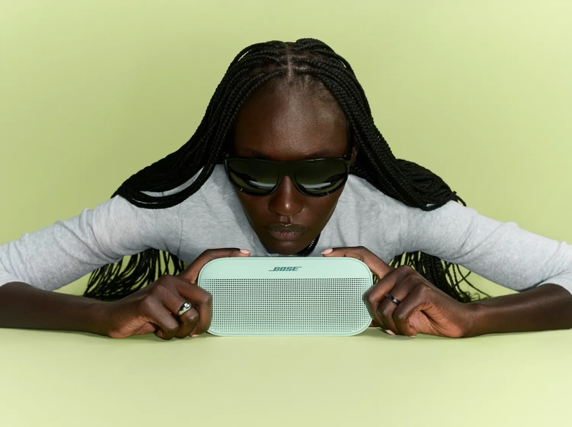 Eine Frau mit Sonnenbrille und Zöpfen hält einen hellgrünen Bose Lautsprecher vor sich.