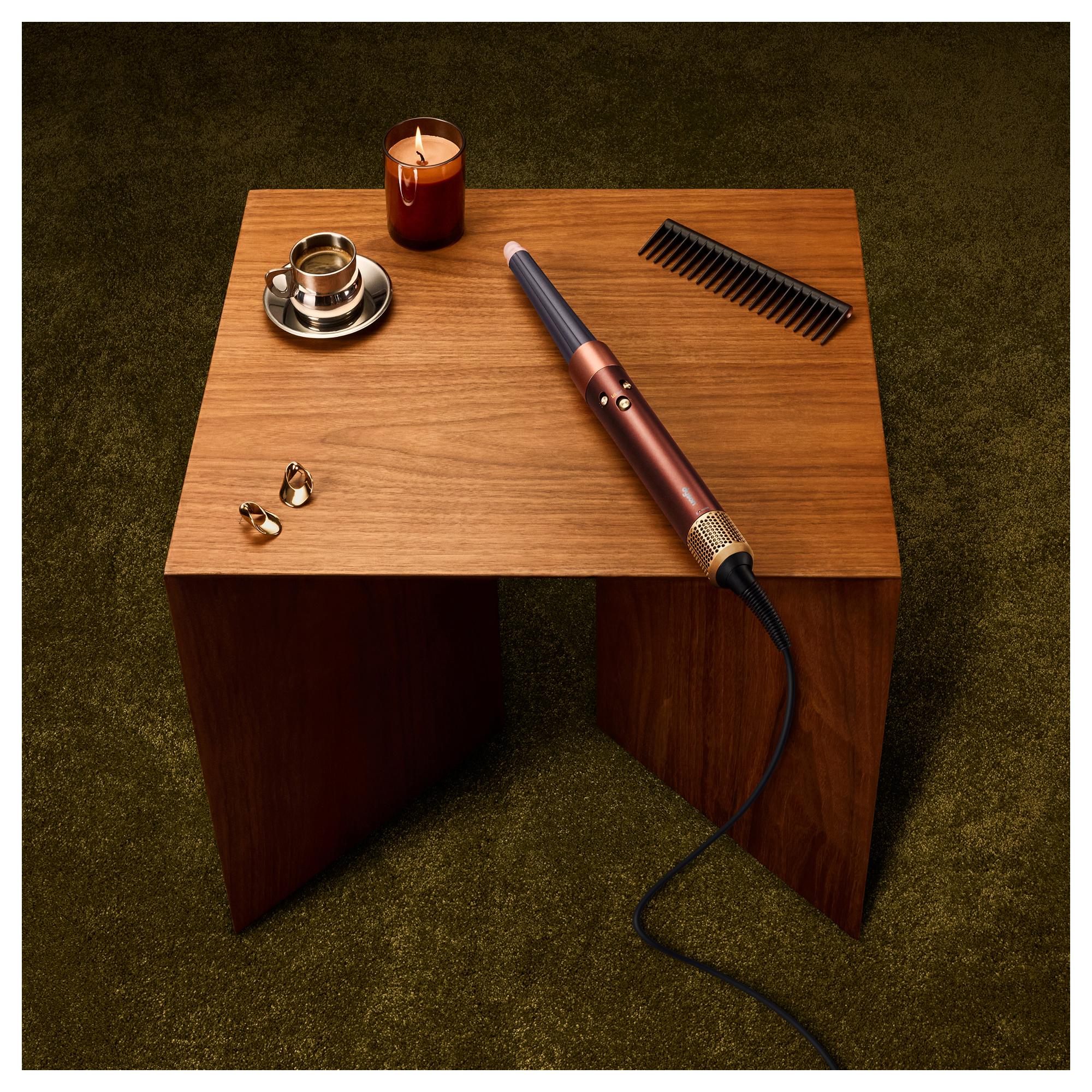 Una mesa de madera en una alfombra verde tiene un rizador, peine, vela, taza de café y aretes.