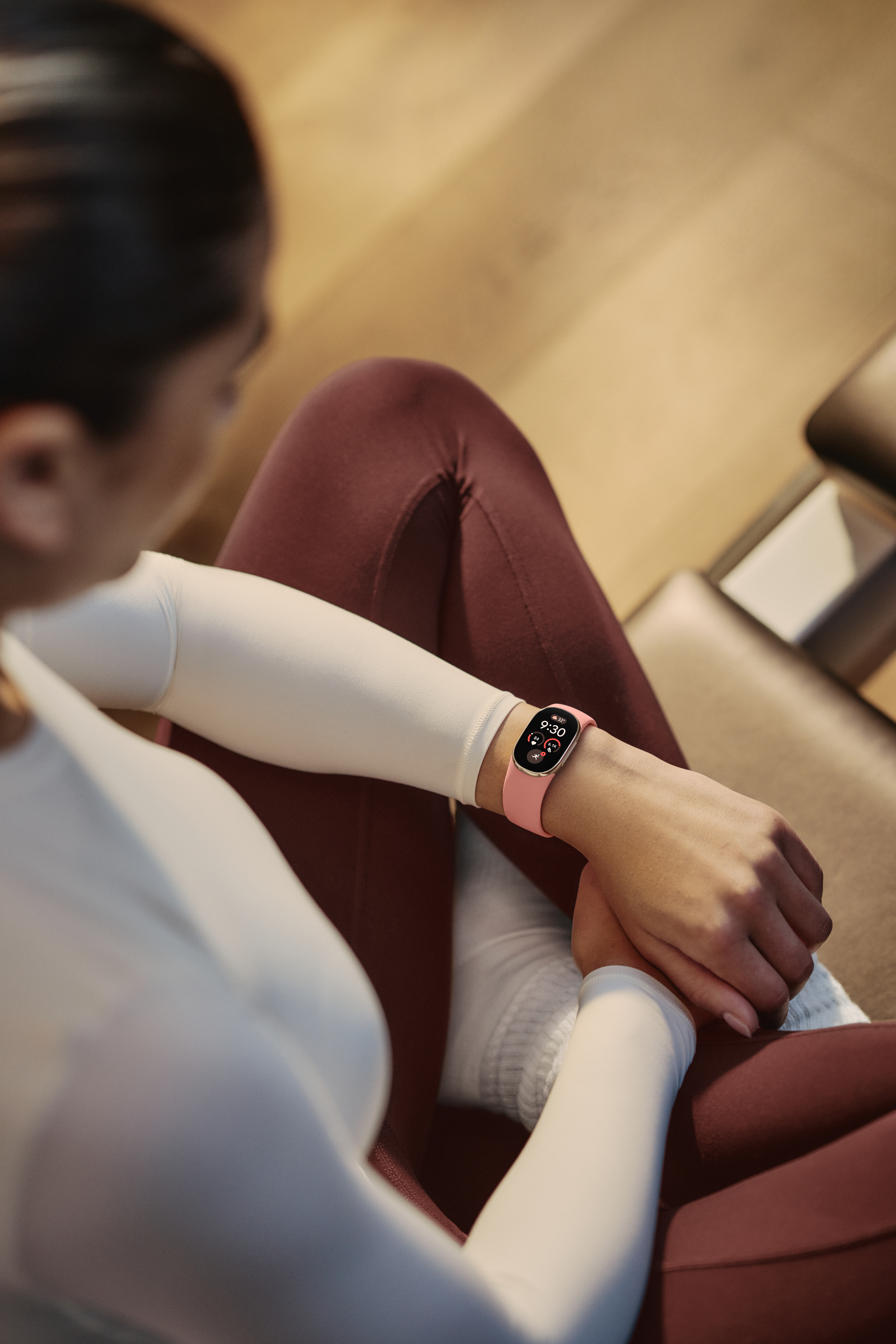 Frau in weißem Oberteil und roten Leggings sitzt und schaut auf ihre rosafarbene Smartwatch.