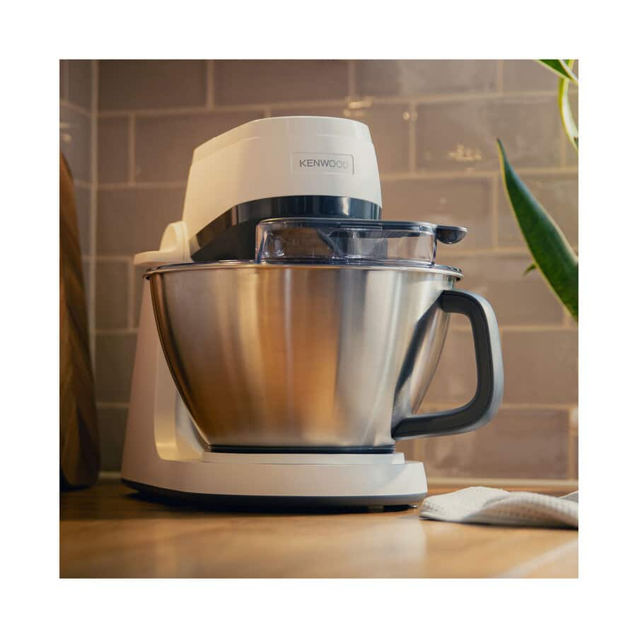 Una batidora blanca Kenwood con bol de acero inoxidable sobre una encimera de madera y pared gris.