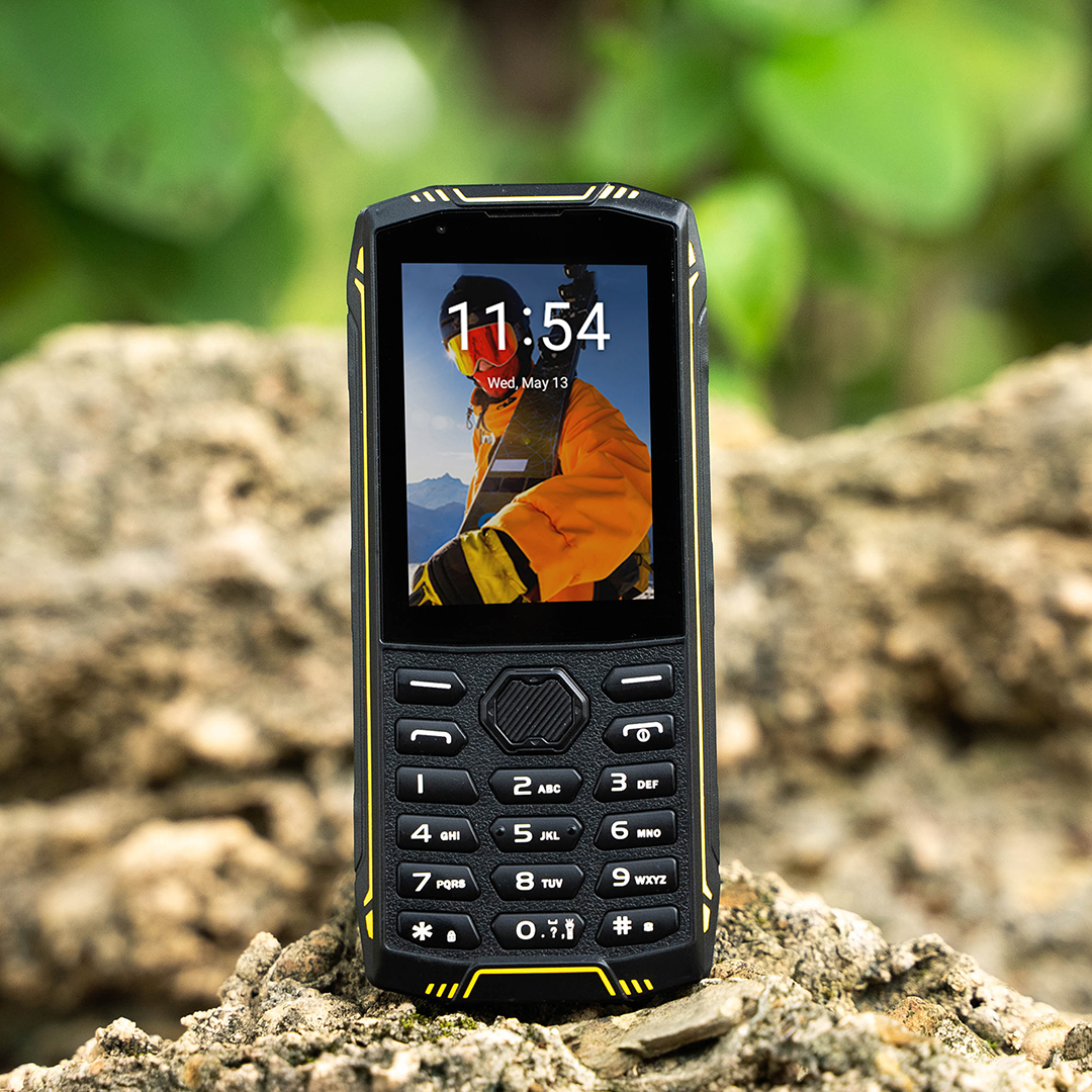 Egy masszív mobiltelefon sárga díszítéssel sziklás felületen áll, háttérben elmosódott zöldséggel.