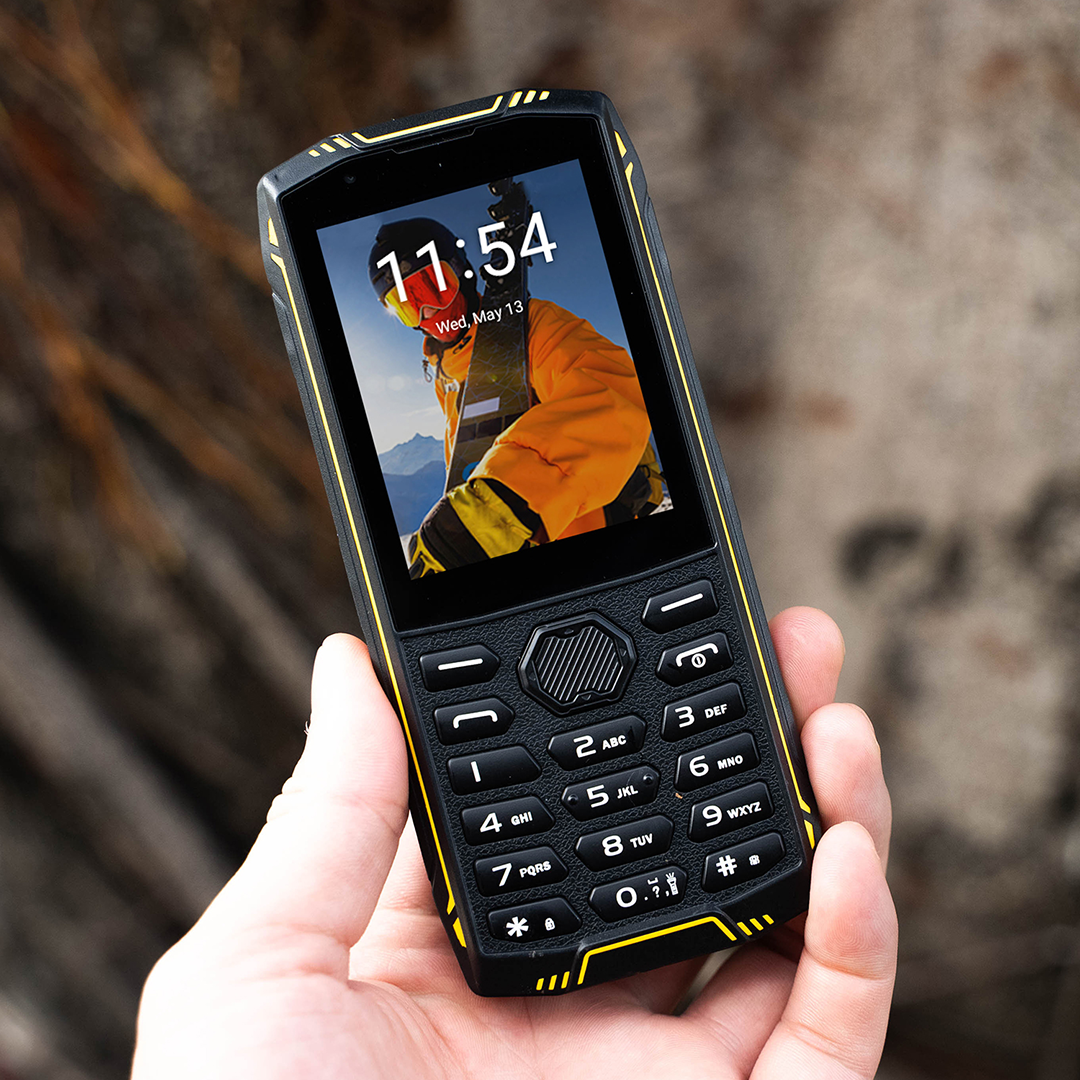 Egy masszív fekete-sárga nyomógombos mobiltelefon kézben tartva, a képernyőn egy síelővel.