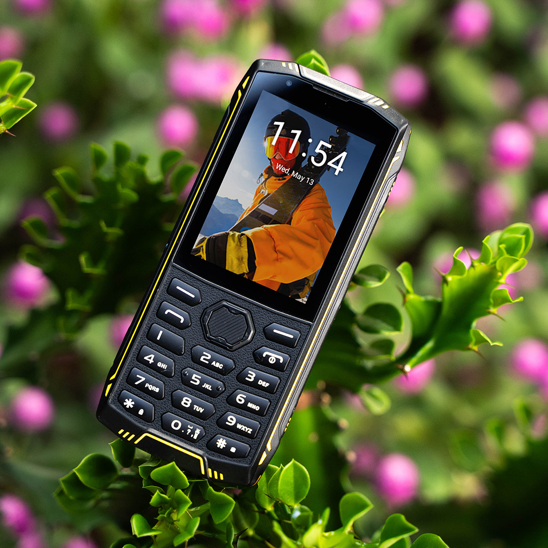 Masszív fekete gombos telefon sárga részletekkel zöld lombozaton rózsaszín virágokkal.