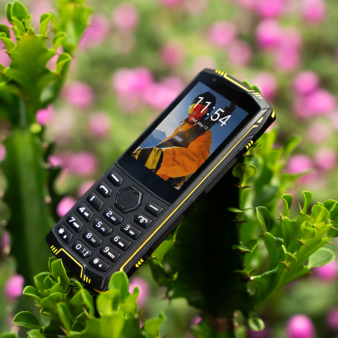 Egy strapabíró fekete-sárga nyomógombos telefon zöld növényen, rózsaszín virágokkal.