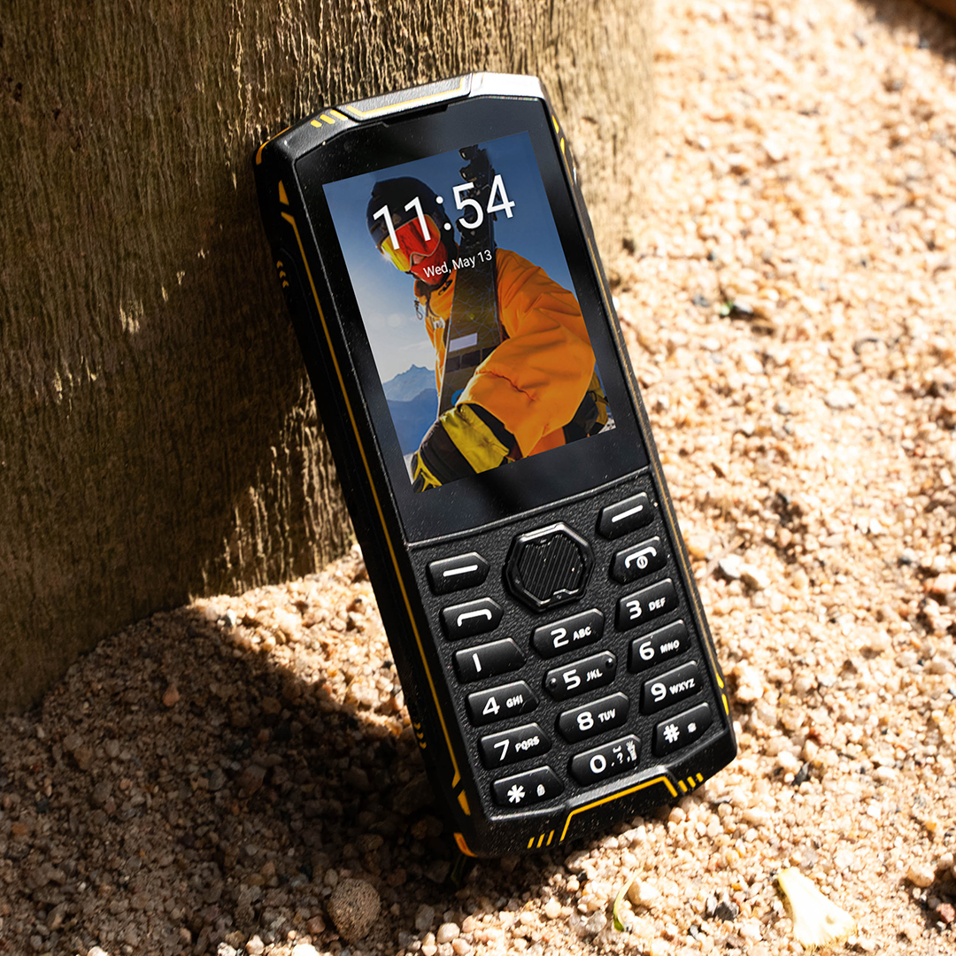 Masszív gombos telefon fizikai billentyűzettel és kijelzővel, fa törzsének támasztva homokos talajon.
