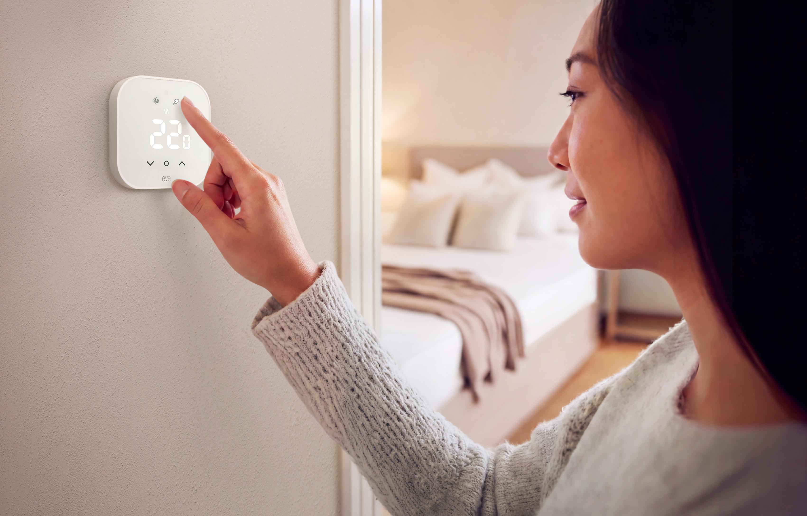 Eine Frau in einem Strickpullover stellt einen weißen Wandthermostat ein, im Hintergrund ist ein Schlafzimmer zu sehen.