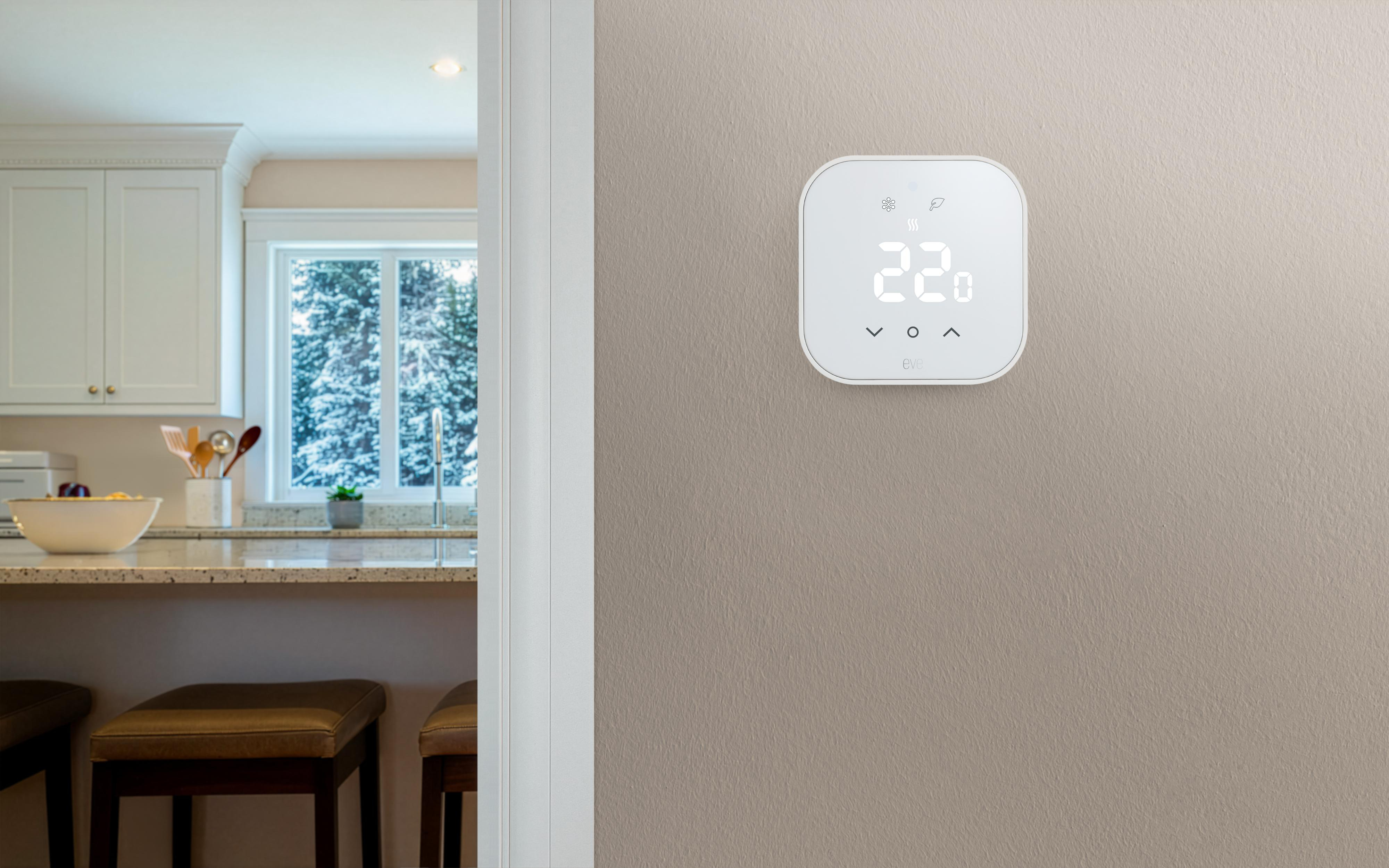 Ein weißer Thermostat an einer grauen Wand zeigt 22 Grad an; im Hintergrund ist eine Küche mit verschneitem Fenster zu sehen.