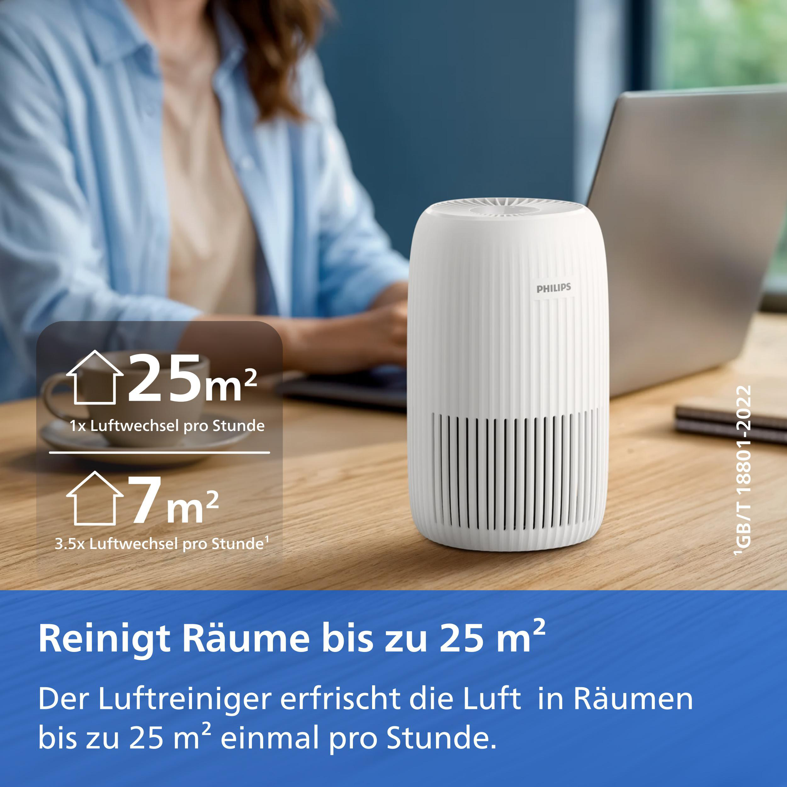 Ein weißer Philips Luftreiniger auf einem Holztisch, im Hintergrund arbeitet eine Frau am Laptop.