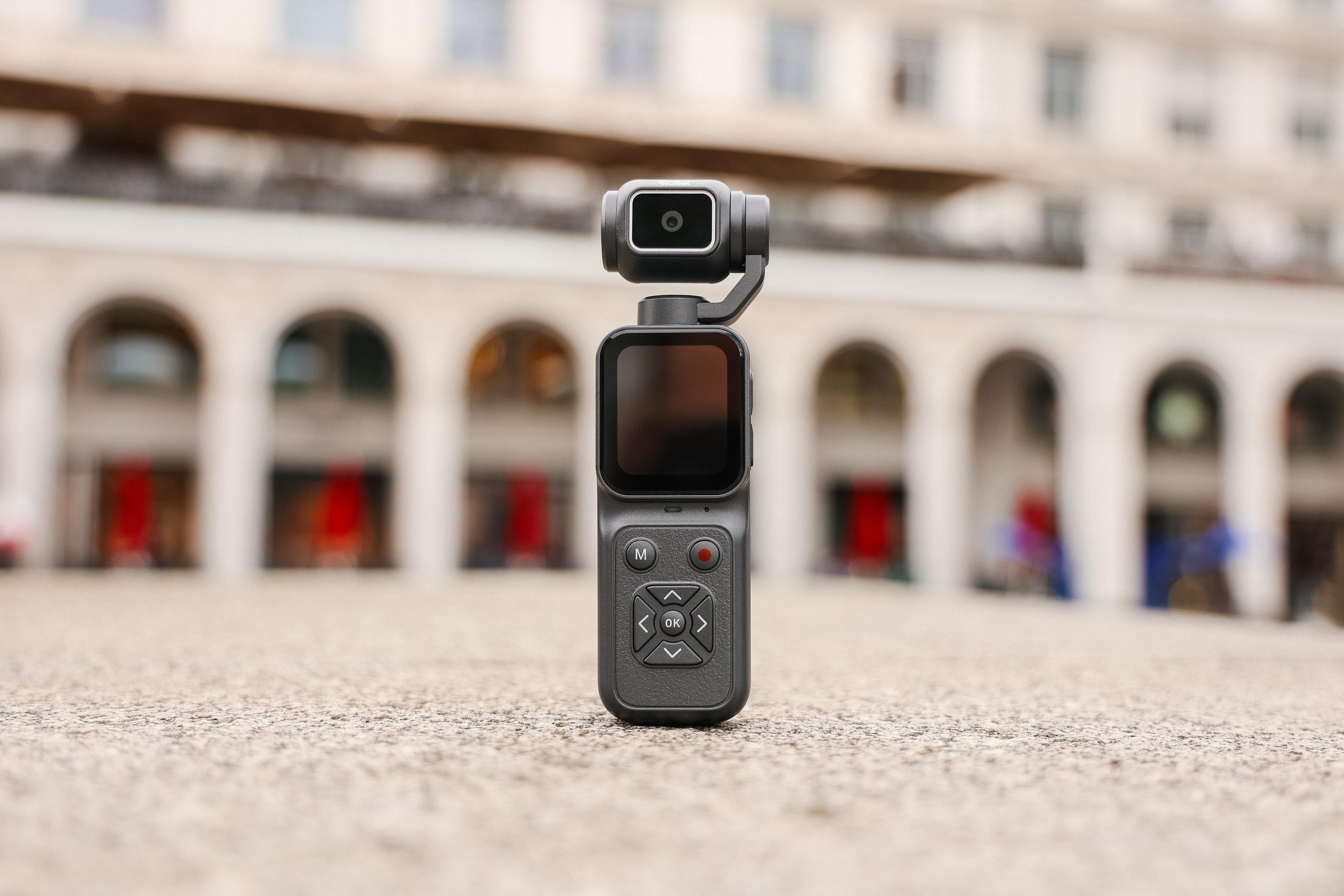 Eine schwarze Handheld-Gimbal-Kamera steht im Vordergrund vor einem unscharfen Gebäude mit Arkaden.