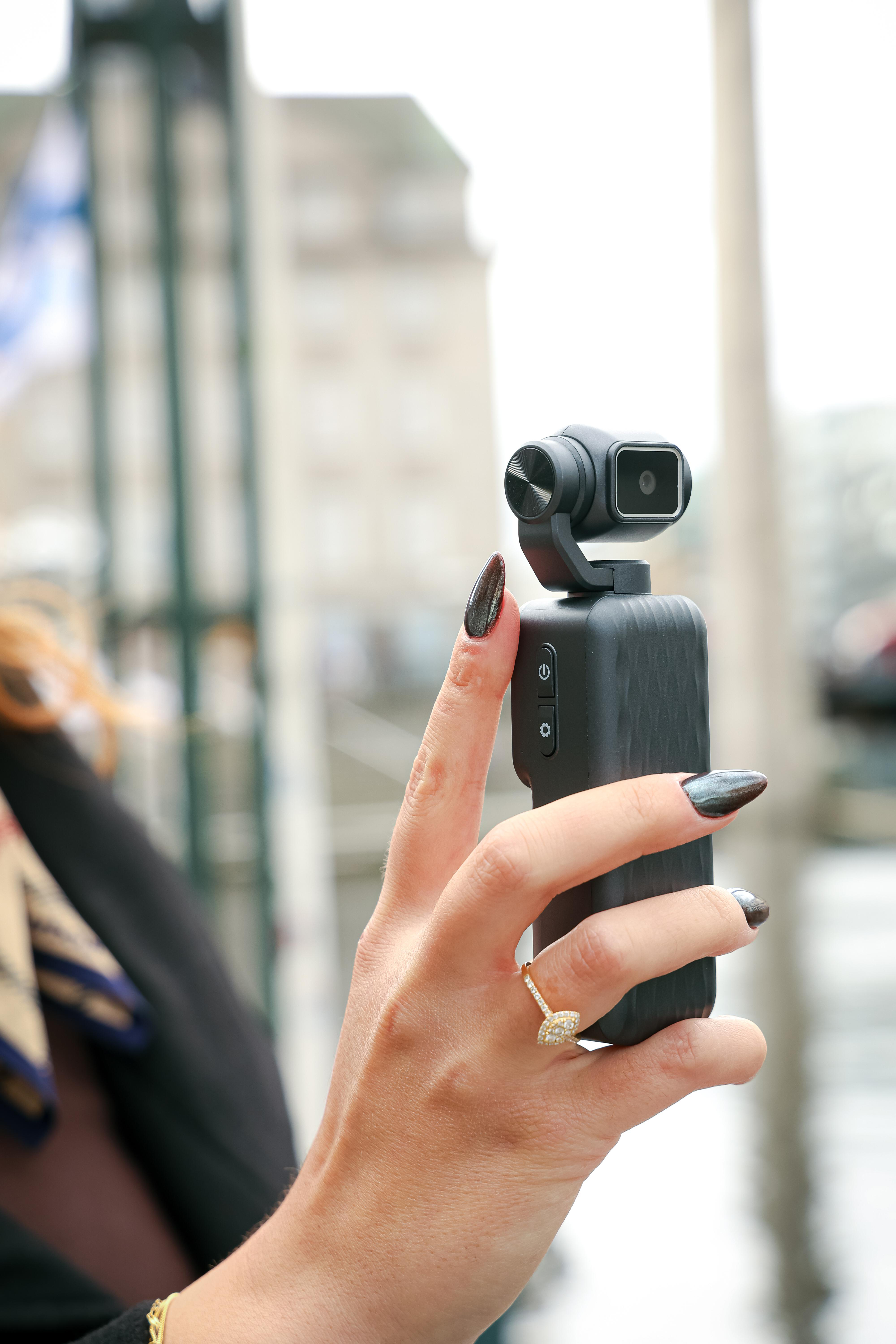Eine Hand mit dunklem Nagellack hält eine kompakte schwarze Videokamera vor einem verschwommenen Hintergrund.