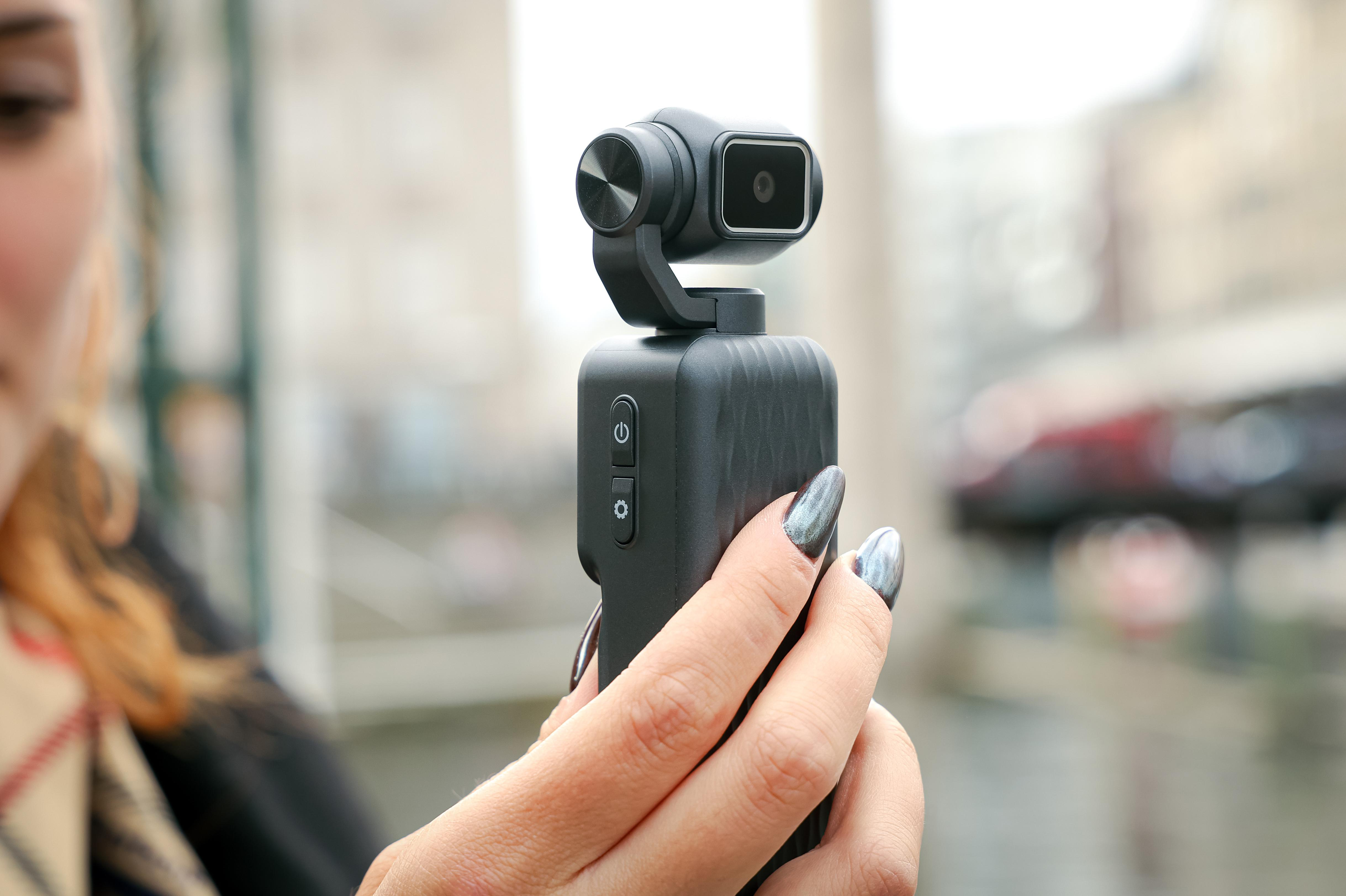 Eine Frau hält eine kompakte, schwarze Gimbal-Kamera mit einem kleinen Bildschirm und Tasten in der Hand.