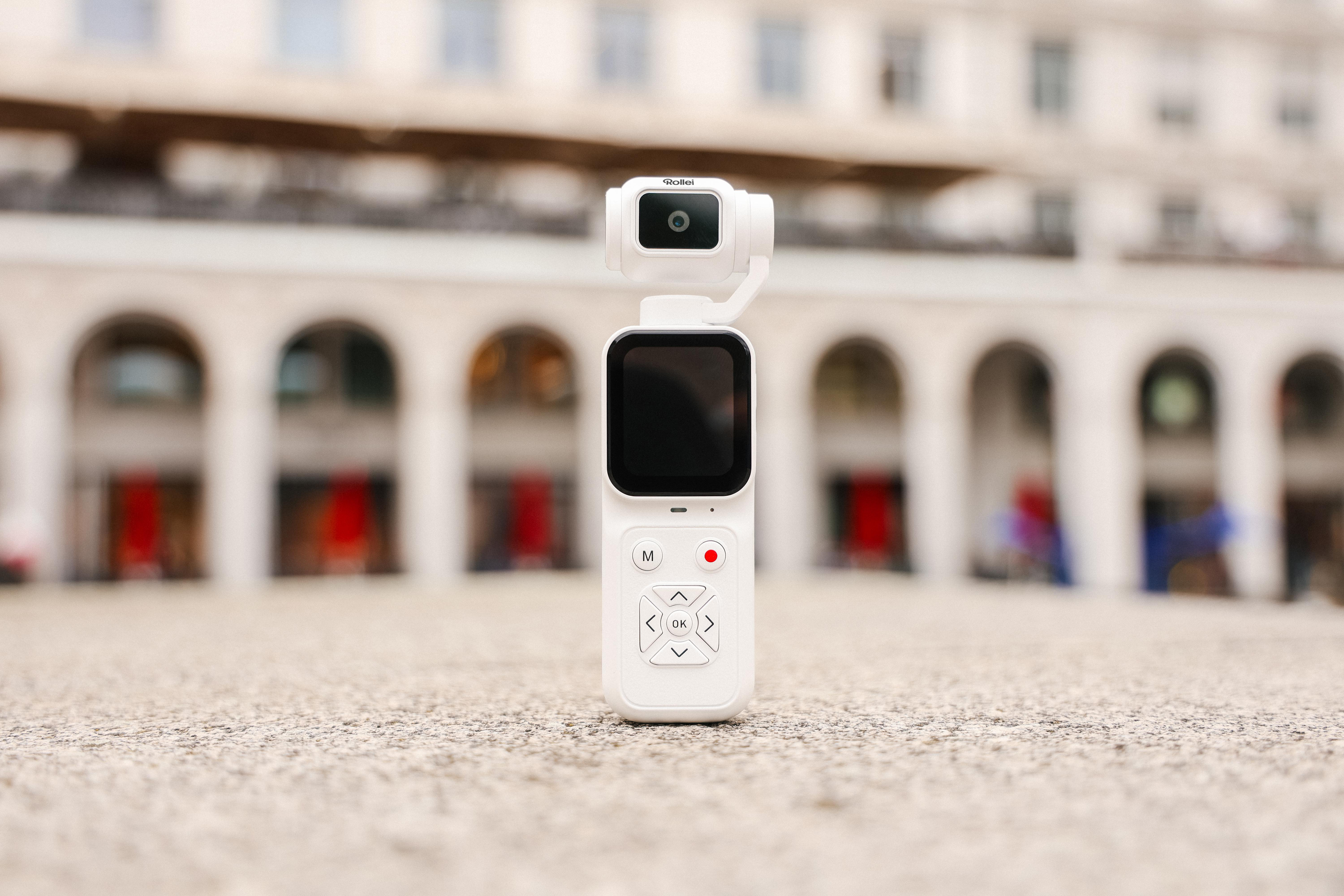 Eine weiße Gimbal-Kamera steht im Vordergrund auf einem Steinboden vor einer Arkadenfassade.