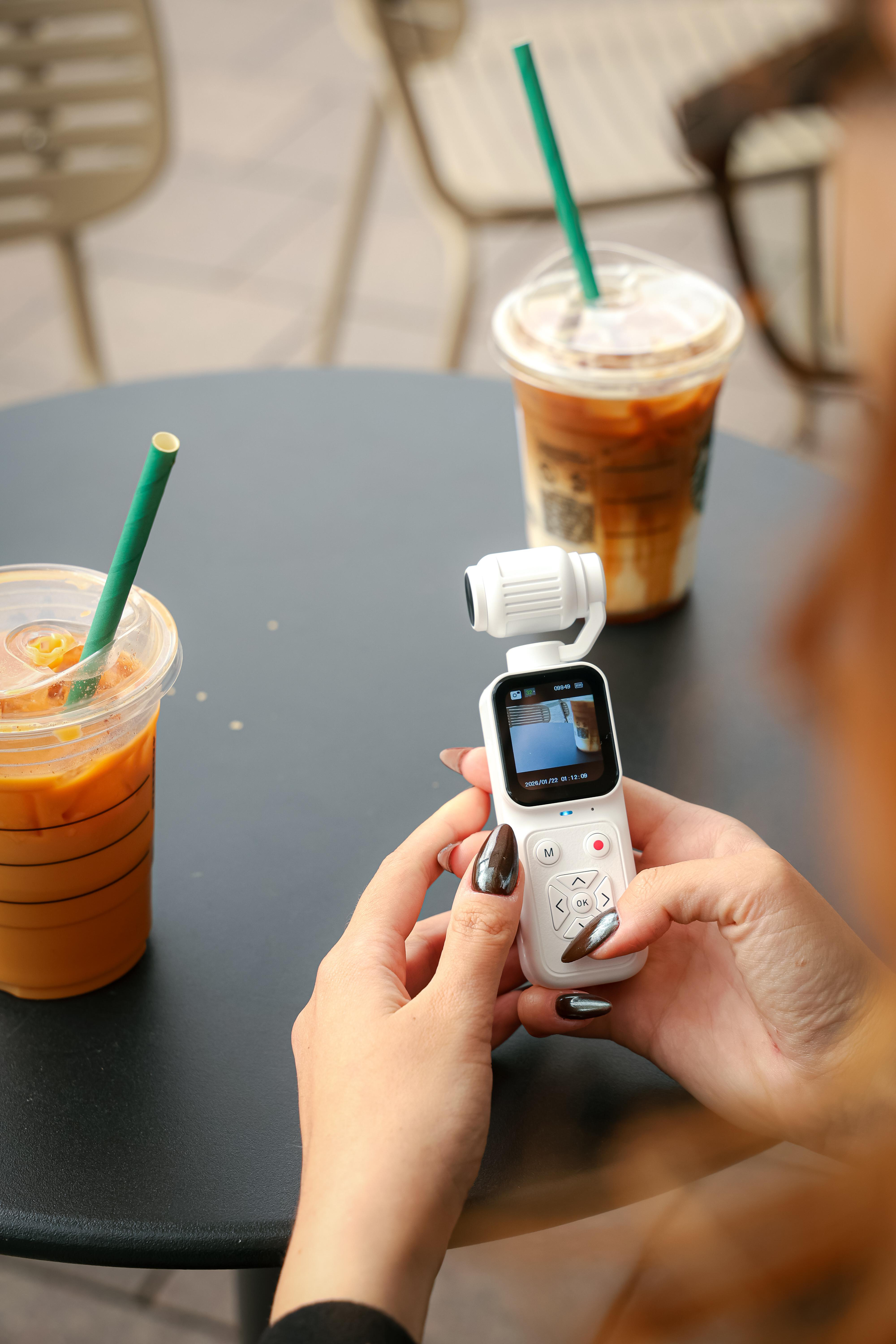 Eine Person hält eine kleine weiße Pocket-Kamera an einem Tisch mit zwei Eiskaffeegetränken.