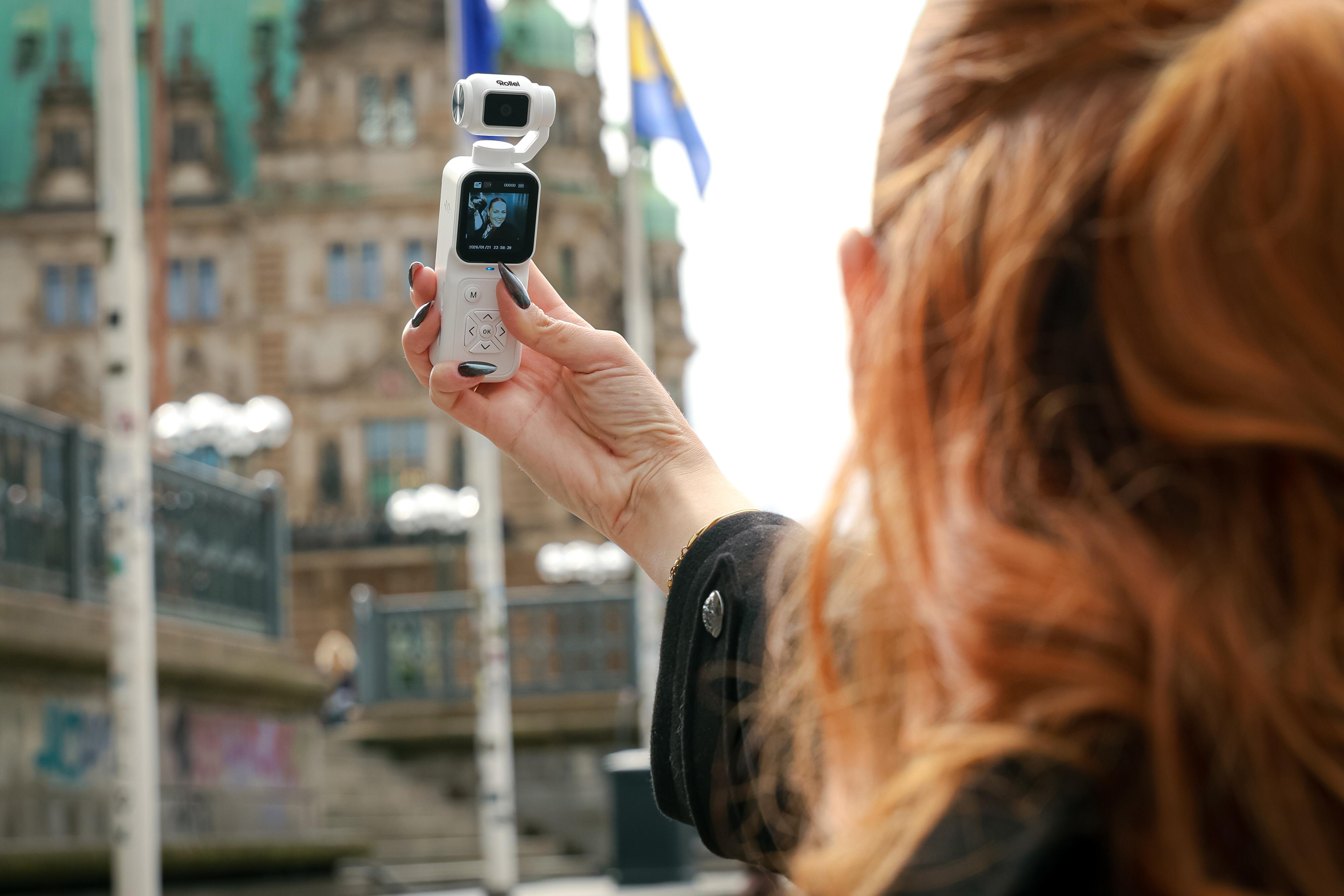 Eine Rothaarige hält vor historischer Kulisse eine weiße Gimbal-Kamera in der Hand.