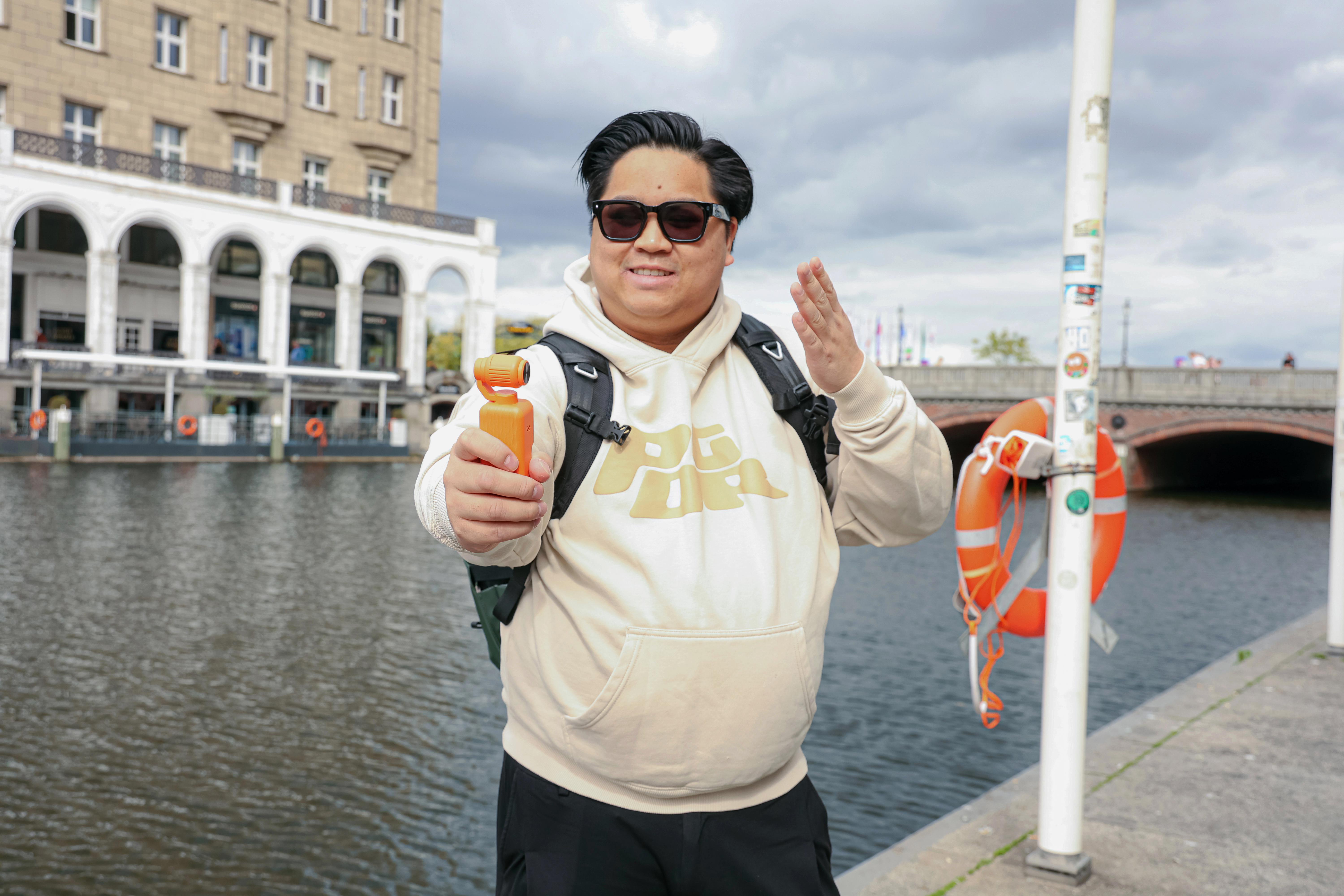 Ein Mann mit Sonnenbrille und hellem Hoodie hält eine kleine Kamera am Kanal.