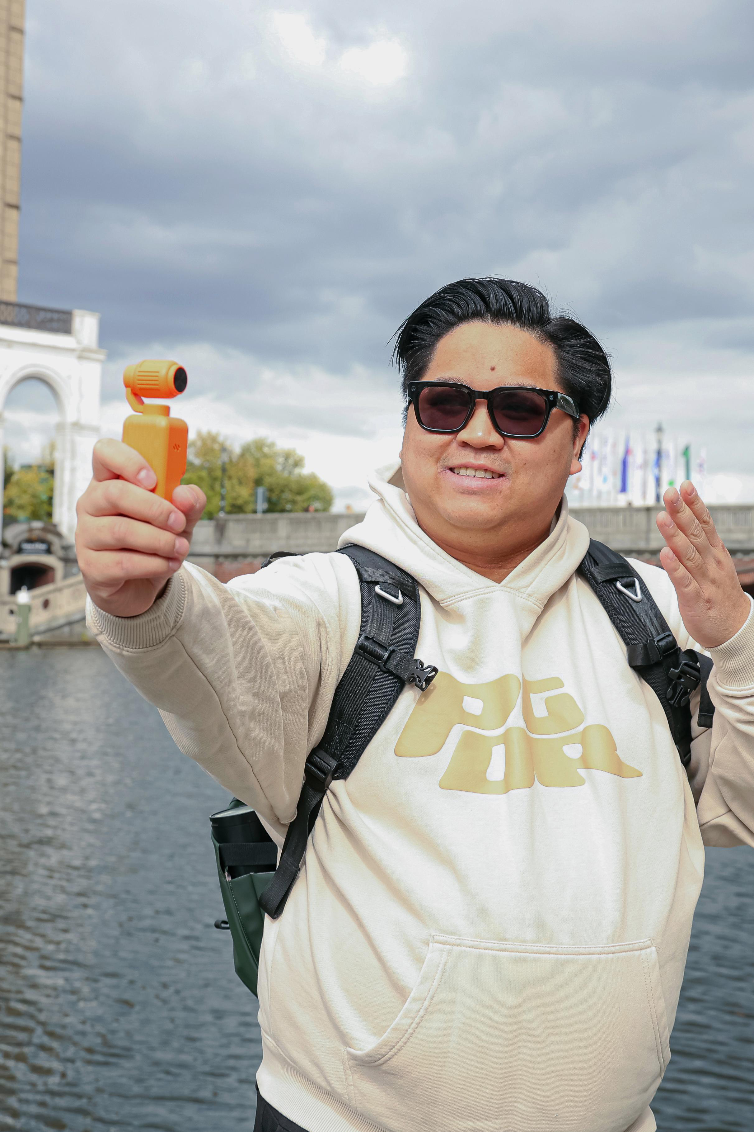 Ein Mann mit Sonnenbrille und Kapuzenpulli hält ein kleines gelbes Gerät für ein Selfie an einem Fluss.