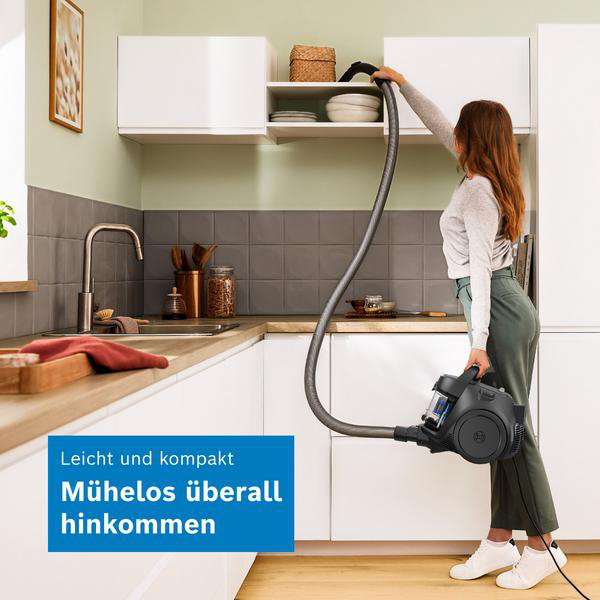 Eine Frau in einer Küche hält einen kompakten Staubsauger und saugt ein oberes Regal ab.