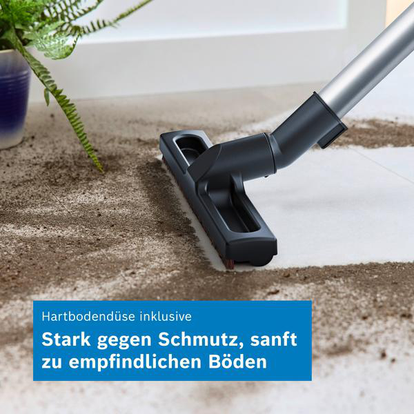 Ein Staubsauger reinigt Schmutz von einem harten Boden, ein grüner Pflanzenfopf steht im Hintergrund.