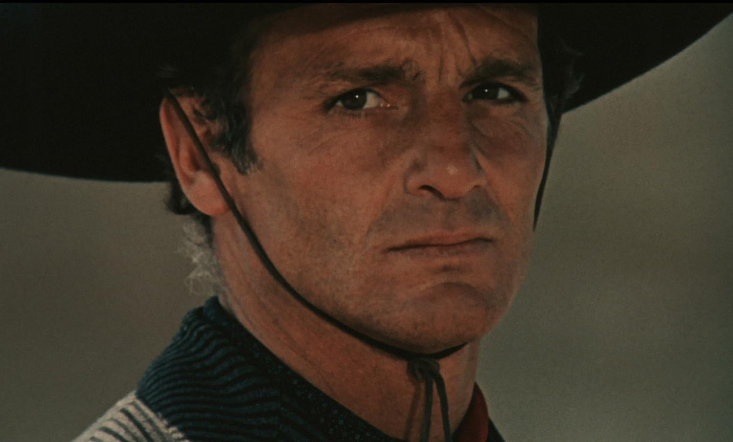 Nahaufnahme eines ernsten Mannes mit dunklem Cowboyhut und gemustertem Poncho, der ernst nach vorne blickt.
