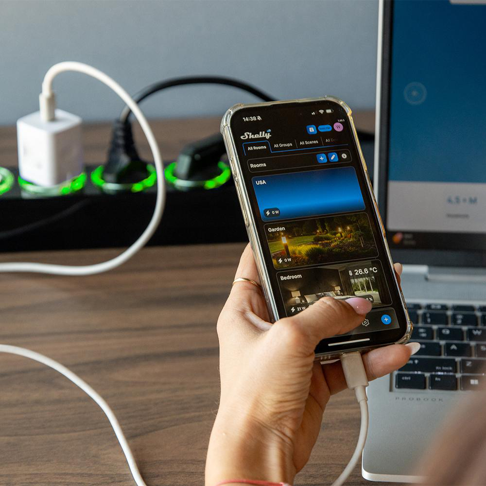 Eine Person hält ein Smartphone mit Smart-Home-App, im Hintergrund Steckdosenleiste und Laptop auf Holztisch.