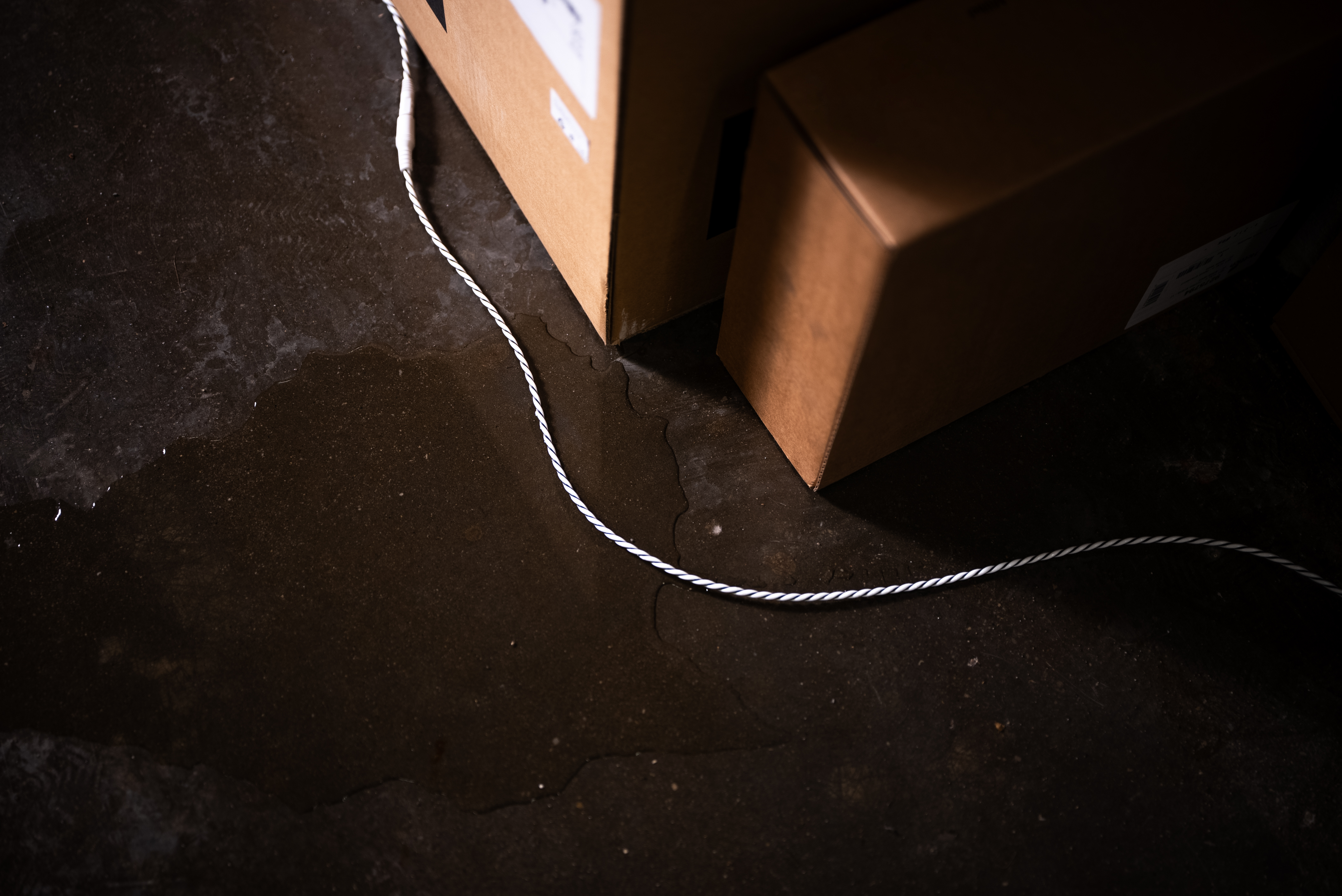 Eine Wasserpfütze auf dem Boden neben braunen Pappkartons und einem weißen Kabel.