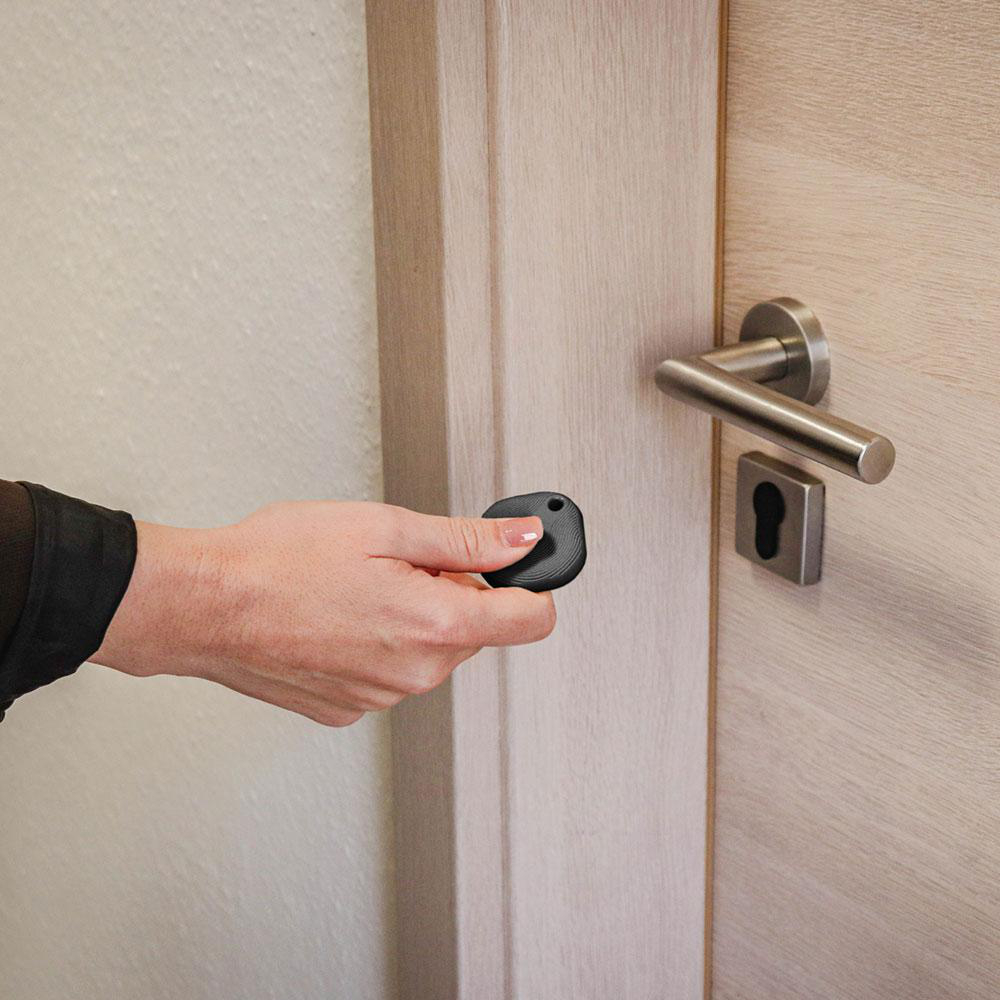 Eine Hand hält einen schwarzen Schlüsselanhänger vor eine Holztür mit Metallgriff.