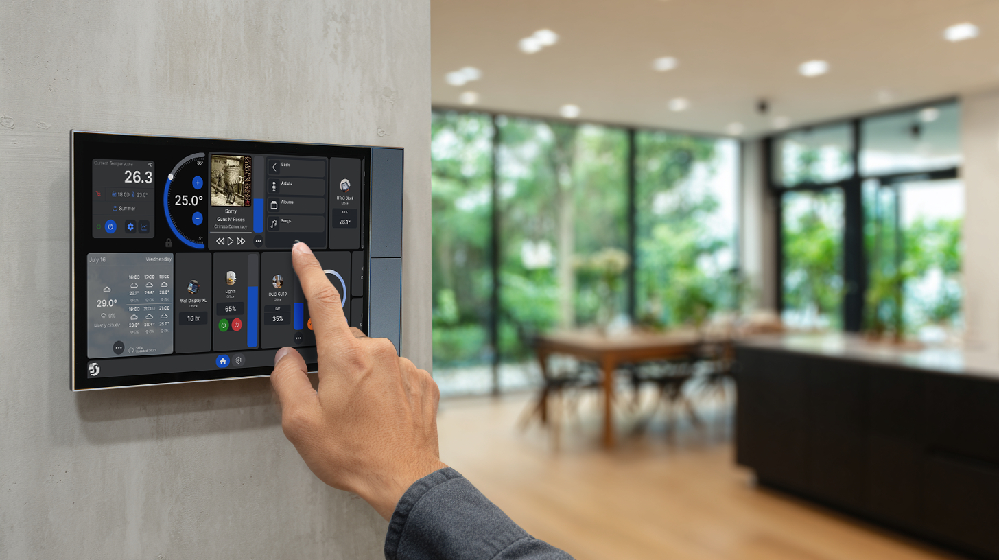 Hand bedient ein schwarzes Smart-Home-Display an einer Wand in einer modernen, offenen Wohnung.