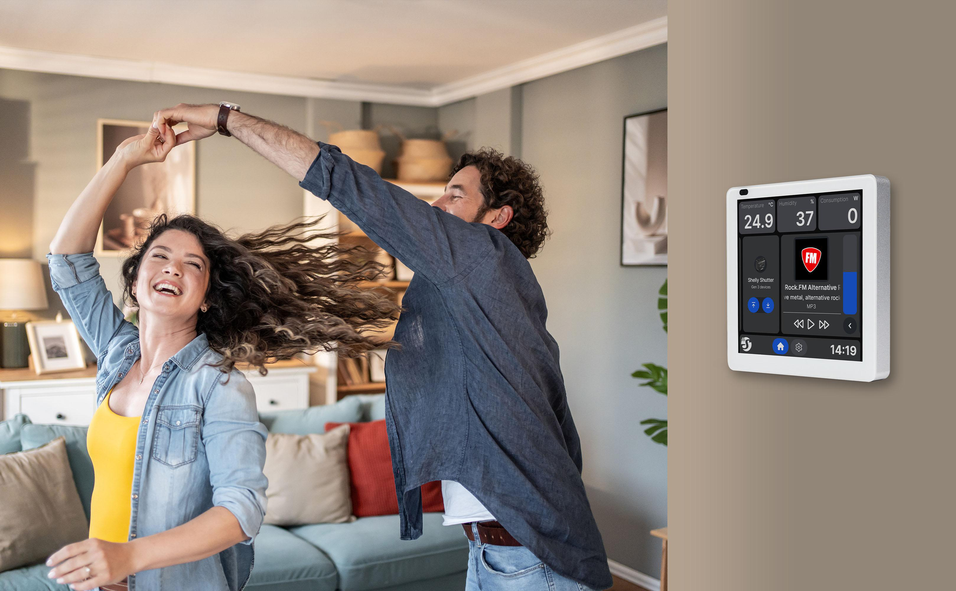 Ein glückliches Paar tanzt im Wohnzimmer, während an der Wand ein digitales Smart-Home-Bedienpanel montiert ist.