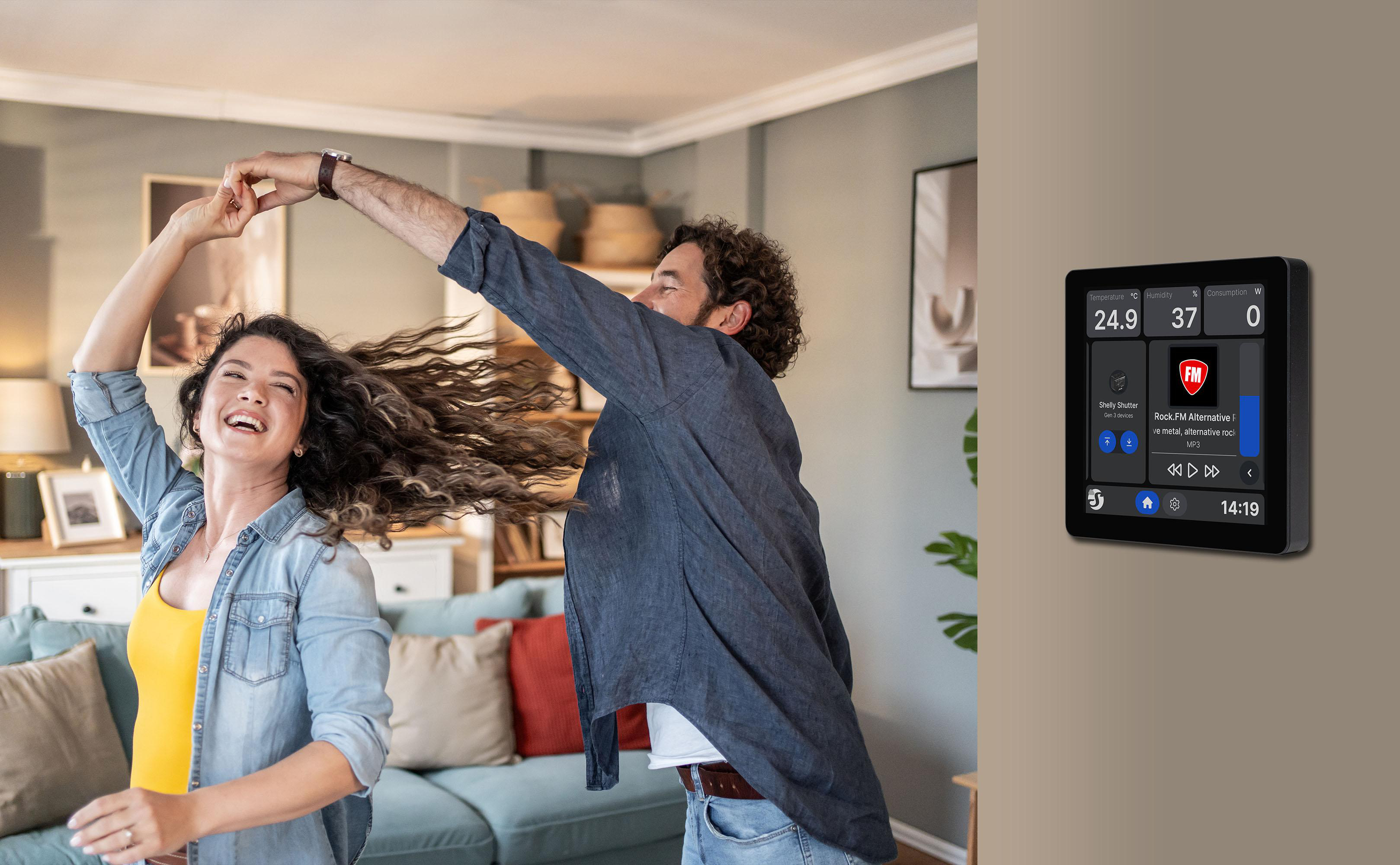 Ein Paar tanzt fröhlich in einem Wohnzimmer neben einem an der Wand montierten Smart-Home-Bedienfeld.