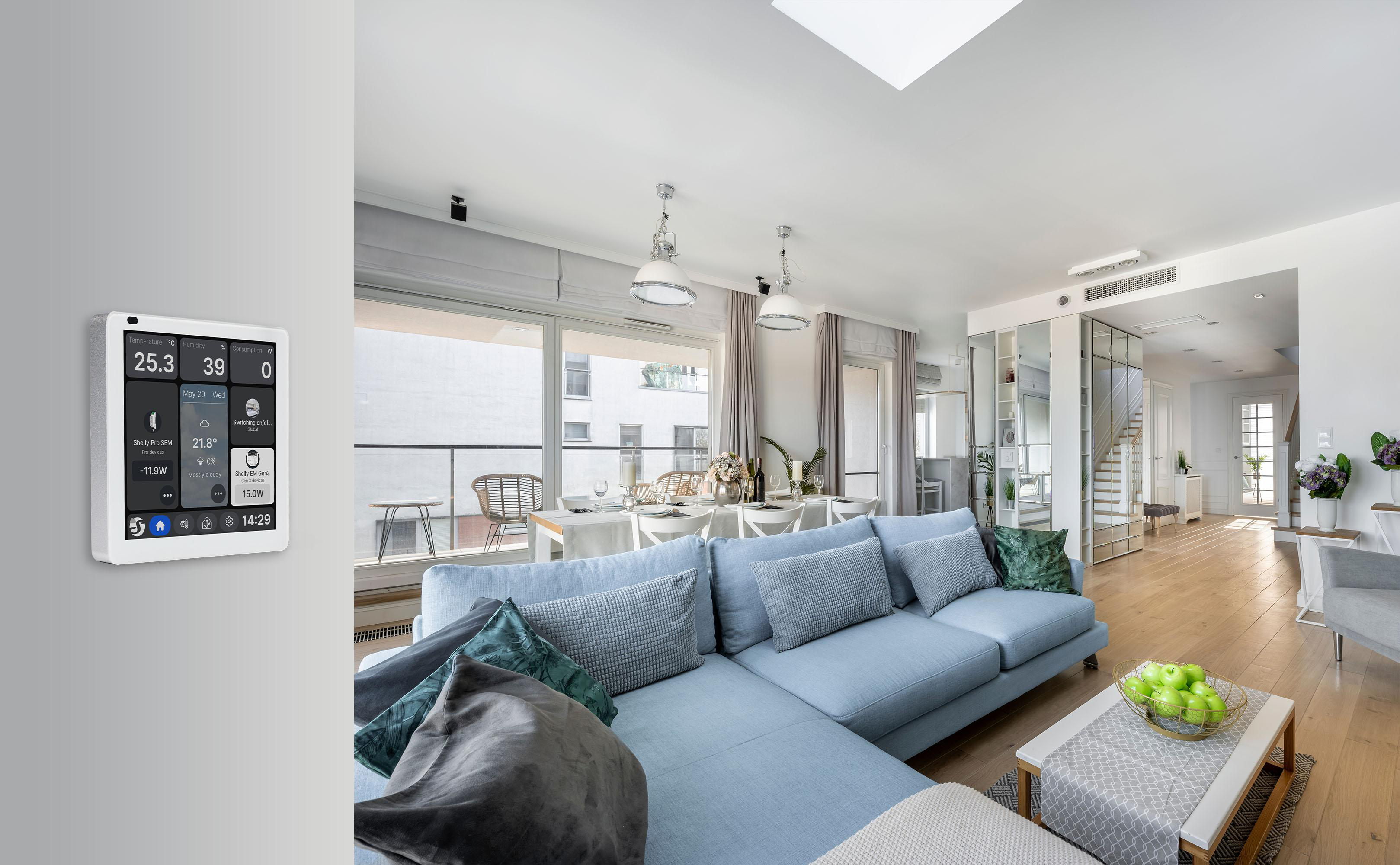Wohnzimmer mit blauem Sofa, weißem Tisch mit grünen Äpfeln und einem digitalen Smart-Home-Display an der Wand.