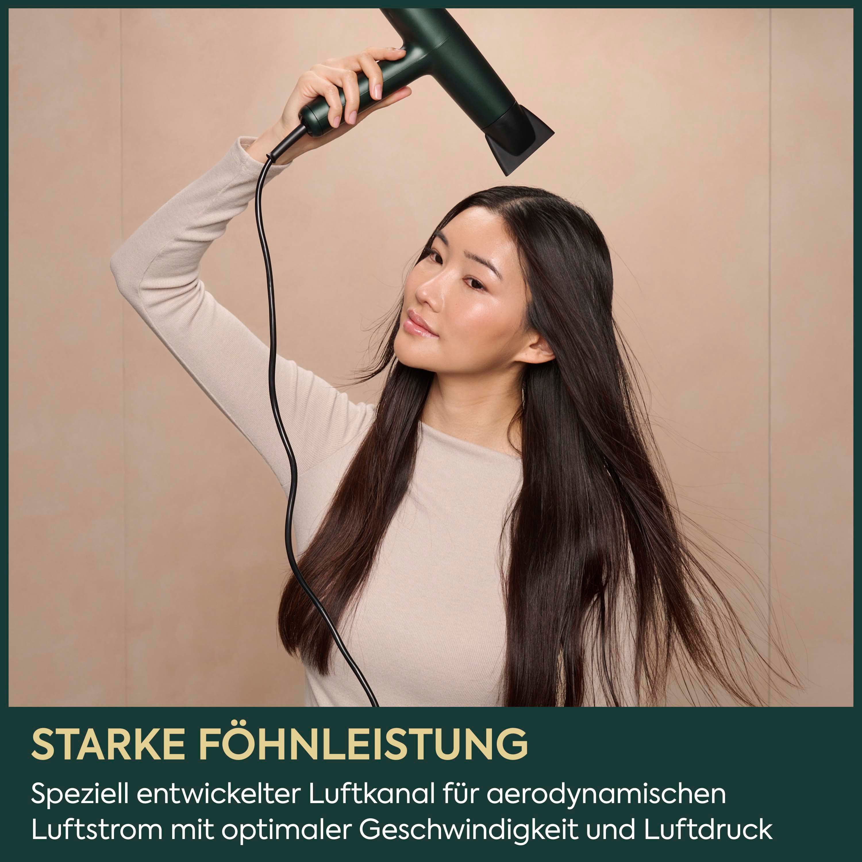 Eine Frau trocknet sich mit einem dunkelgrünen Haartrockner die langen, dunklen Haare vor einem neutralen Hintergrund.
