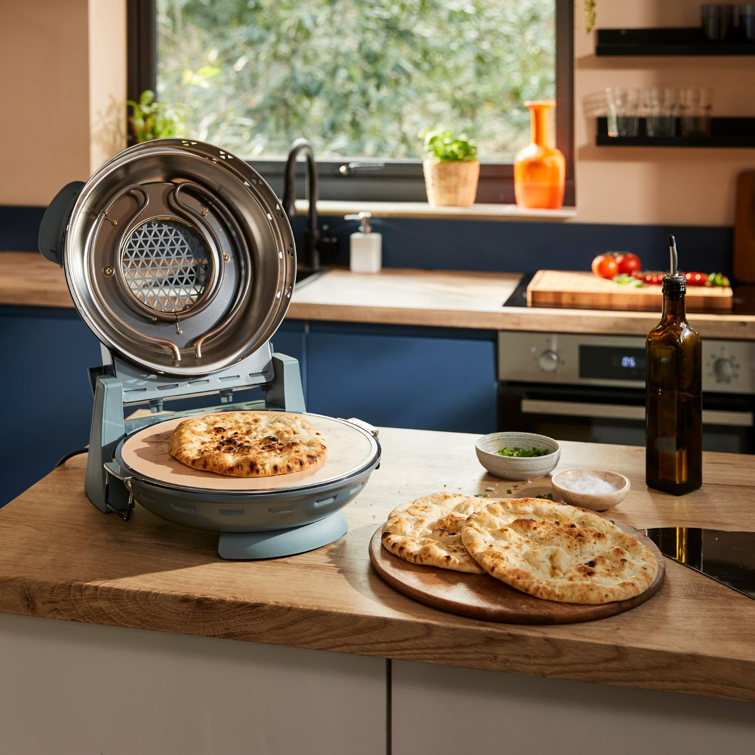 Un horno de pizza eléctrico abierto en una encimera de madera con pan plano cerca.