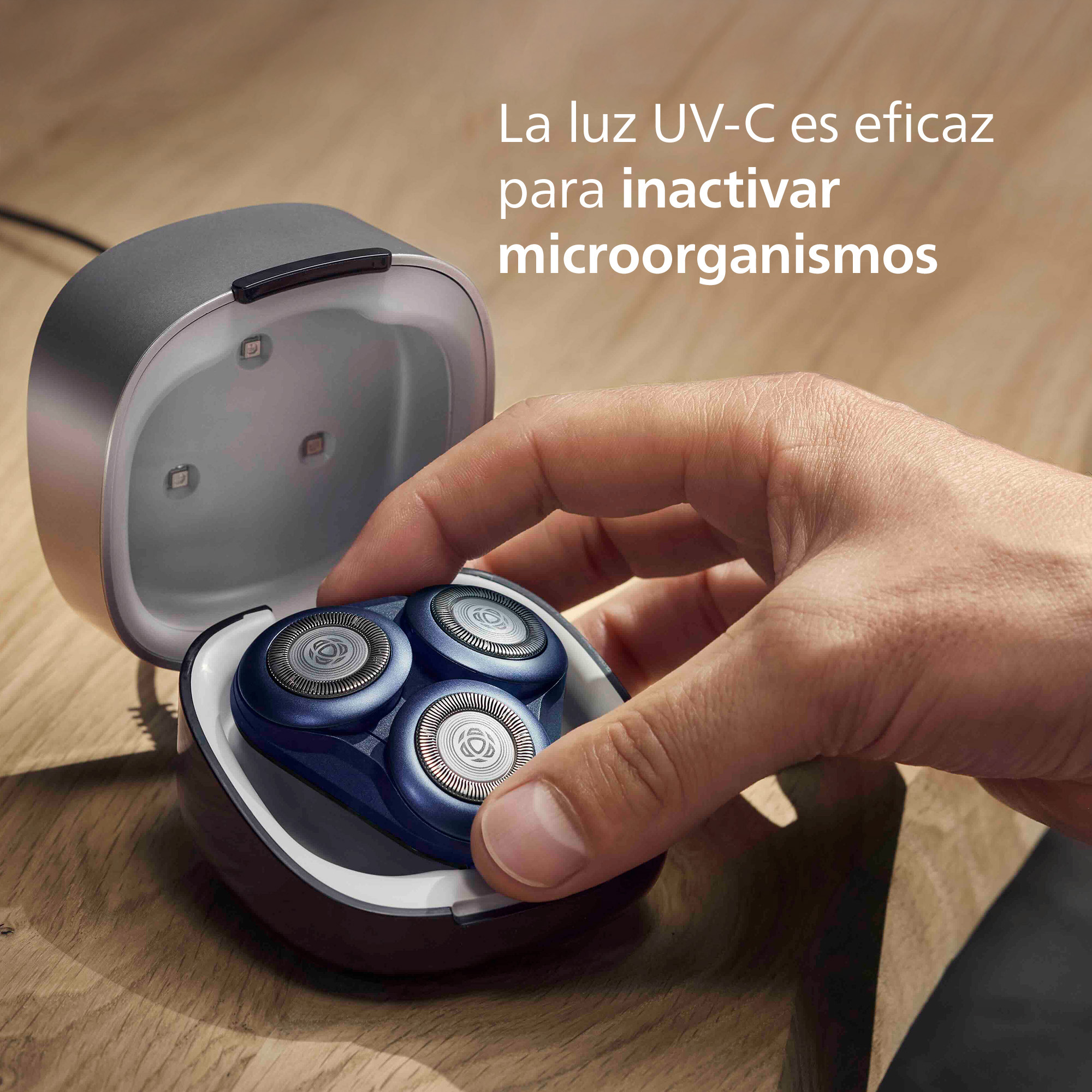 Una mano coloca un cabezal de afeitadora azul en un dispositivo de limpieza UV-C sobre madera.