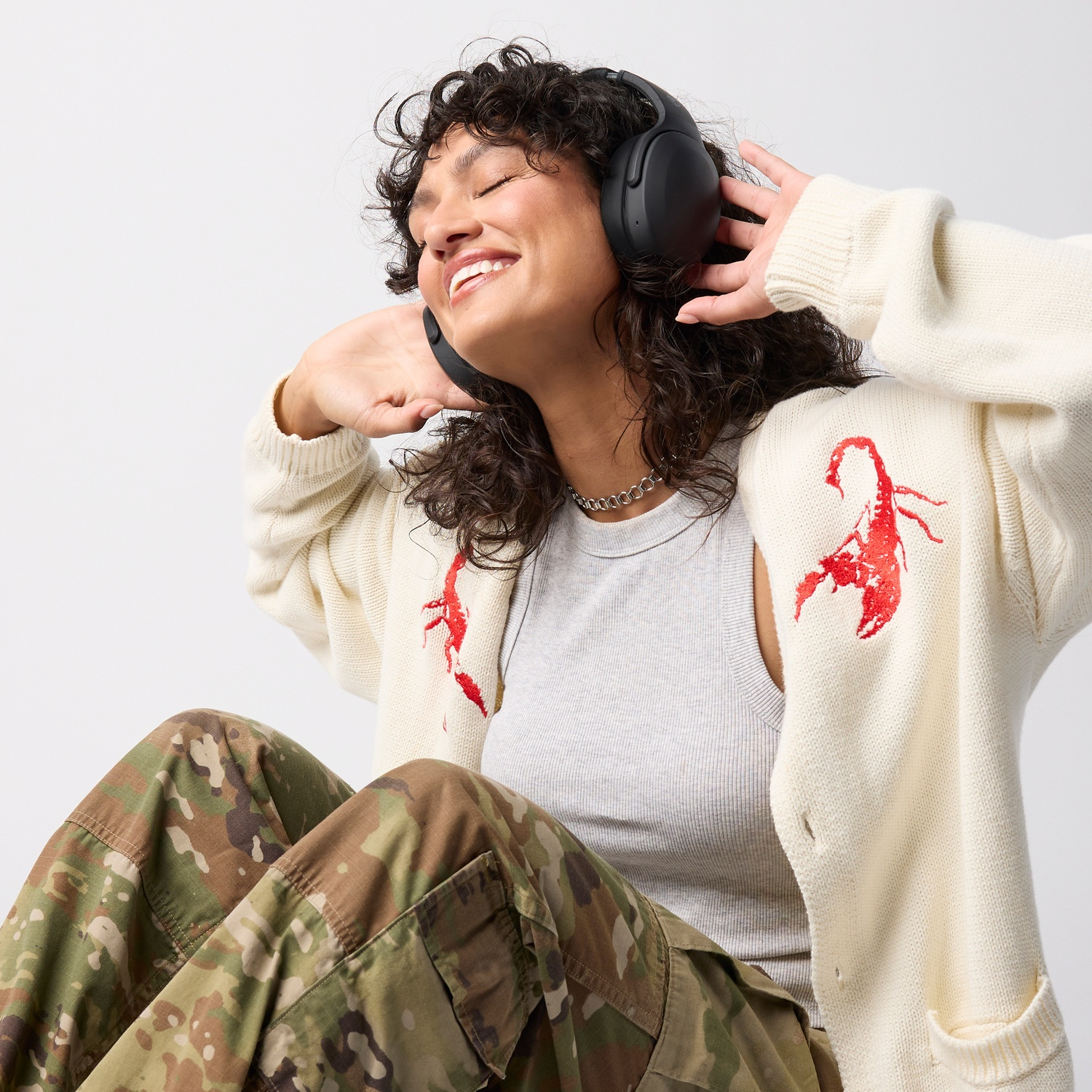 Een vrouw met donker krullend haar, koptelefoon, wit vest met schorpioenborduursel en camouflagebroek lacht.