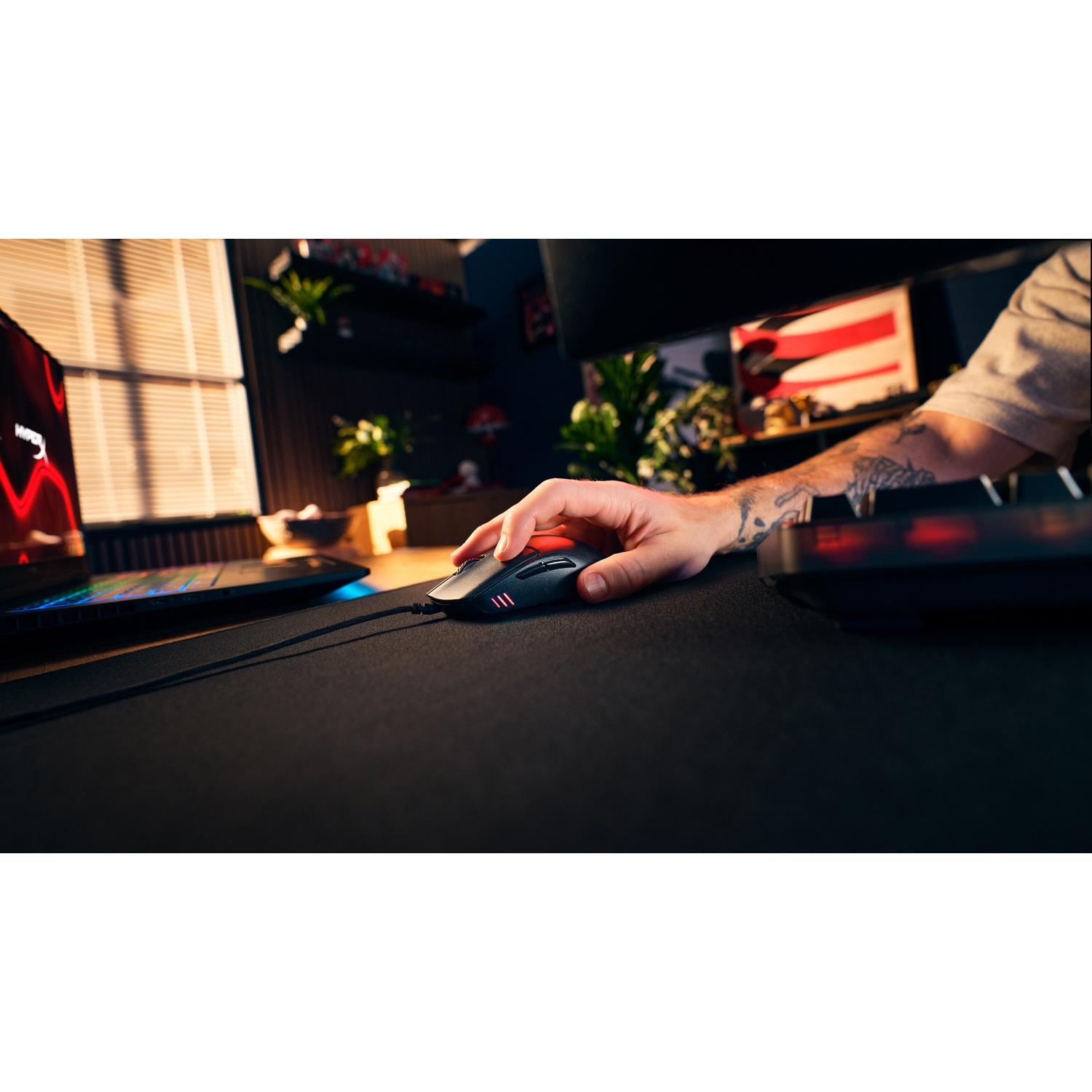 Une main tatouée manipule une souris de jeu filaire noire sur un tapis dans une pièce éclairée.