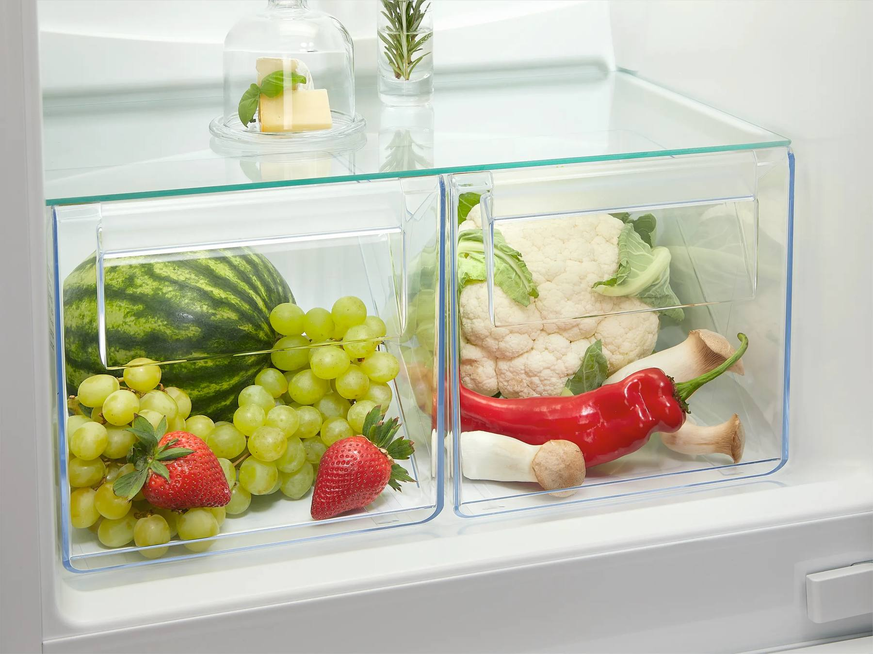 Cajones de refrigerador llenos de frutas frescas como sandía y uvas a la izquierda, y verduras a la derecha.
