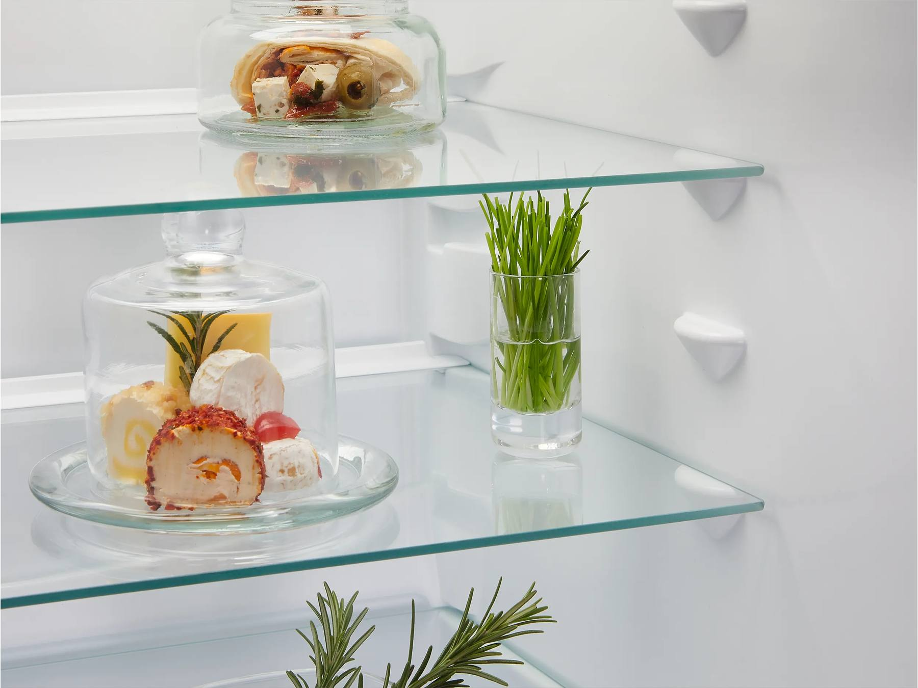 Un refrigerador blanco con estantes de vidrio contiene comida en conserva, queso bajo una cúpula y hierbas frescas.