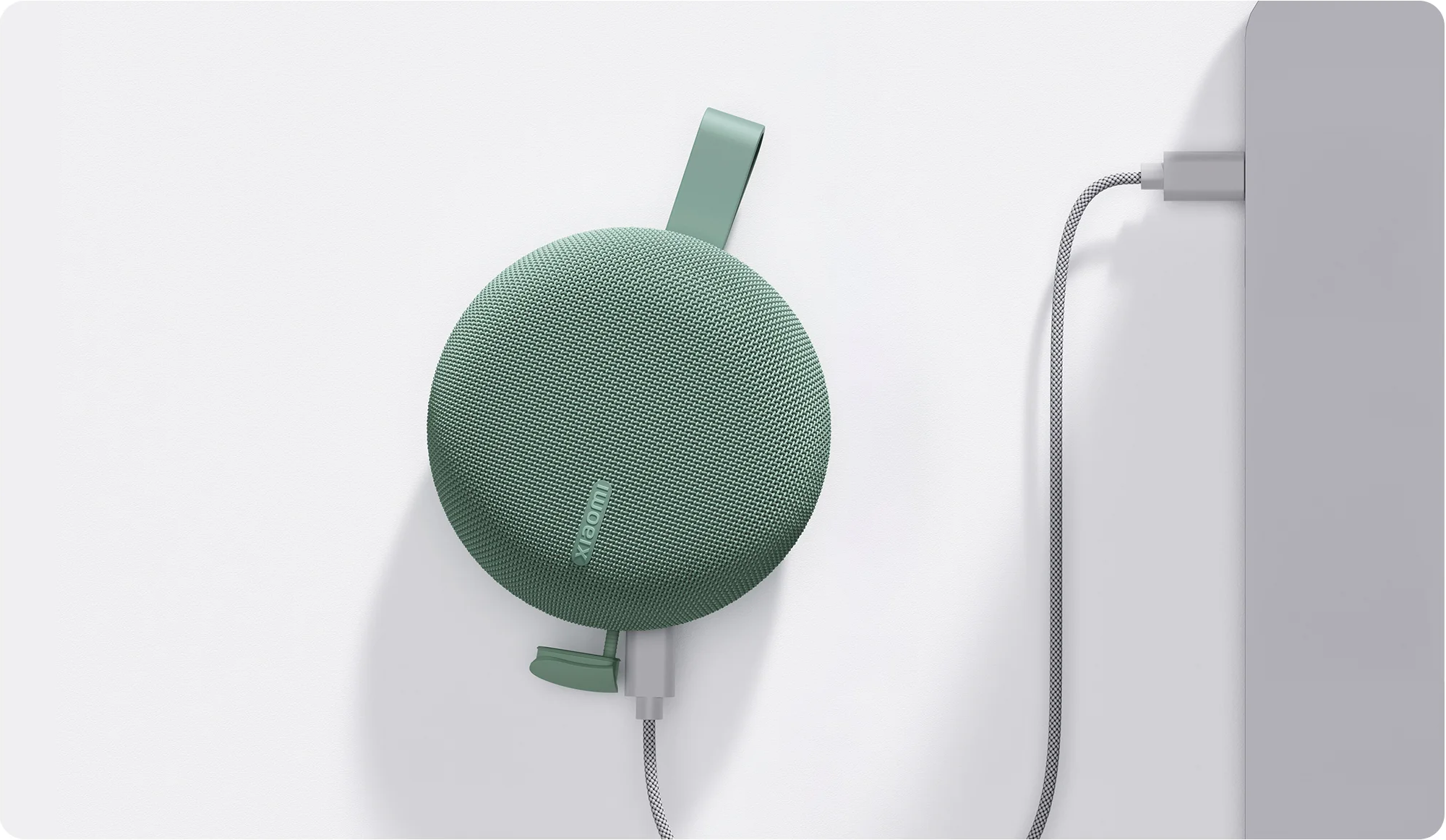 Egy kerek, zöld Xiaomi hangszóró szürke kábellel csatlakozik egy elektronikus eszközhöz.