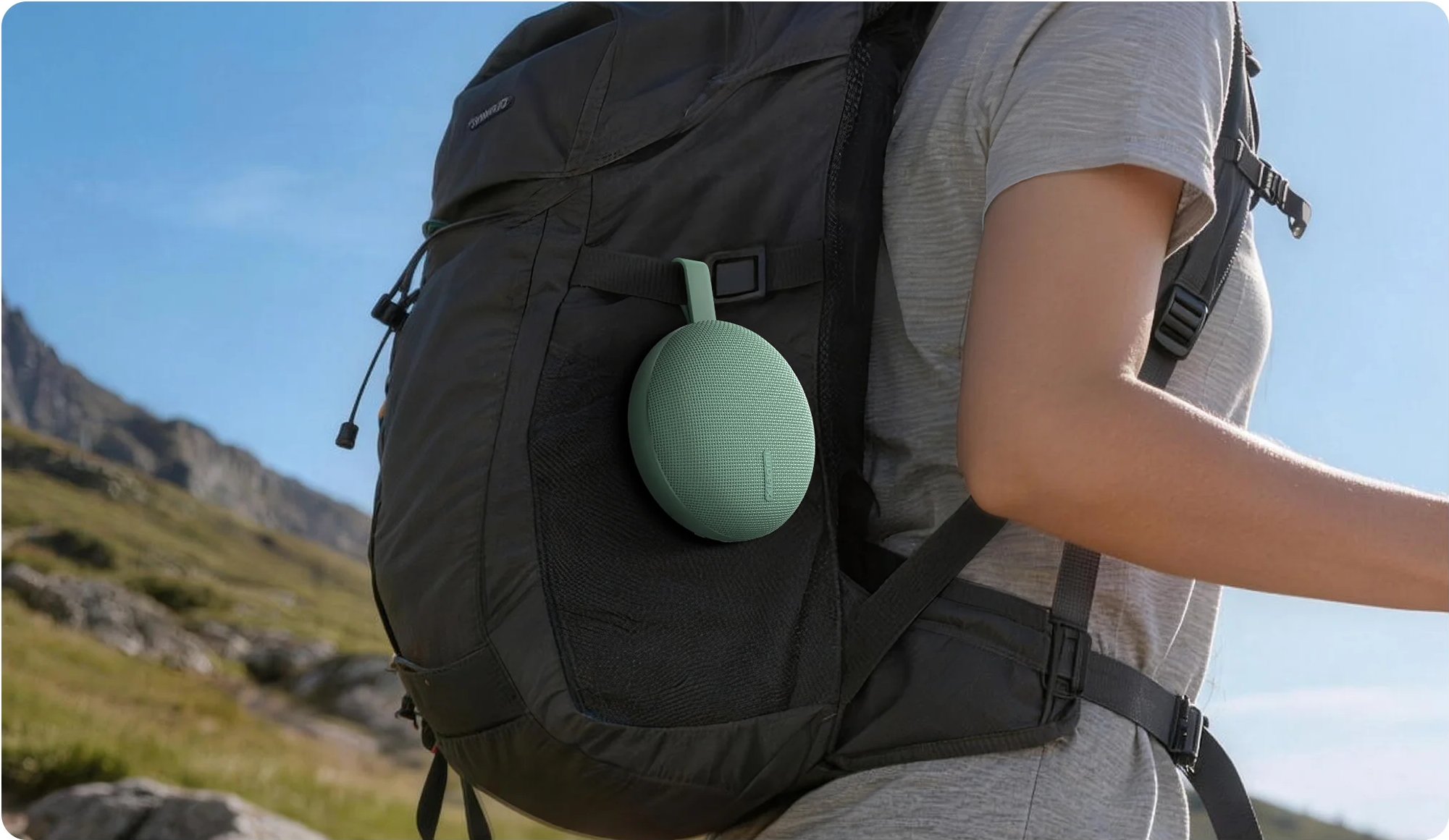 Egy zöld Bluetooth-hangszóró lóg egy fekete hátizsákon egy hegyi táj előtt.