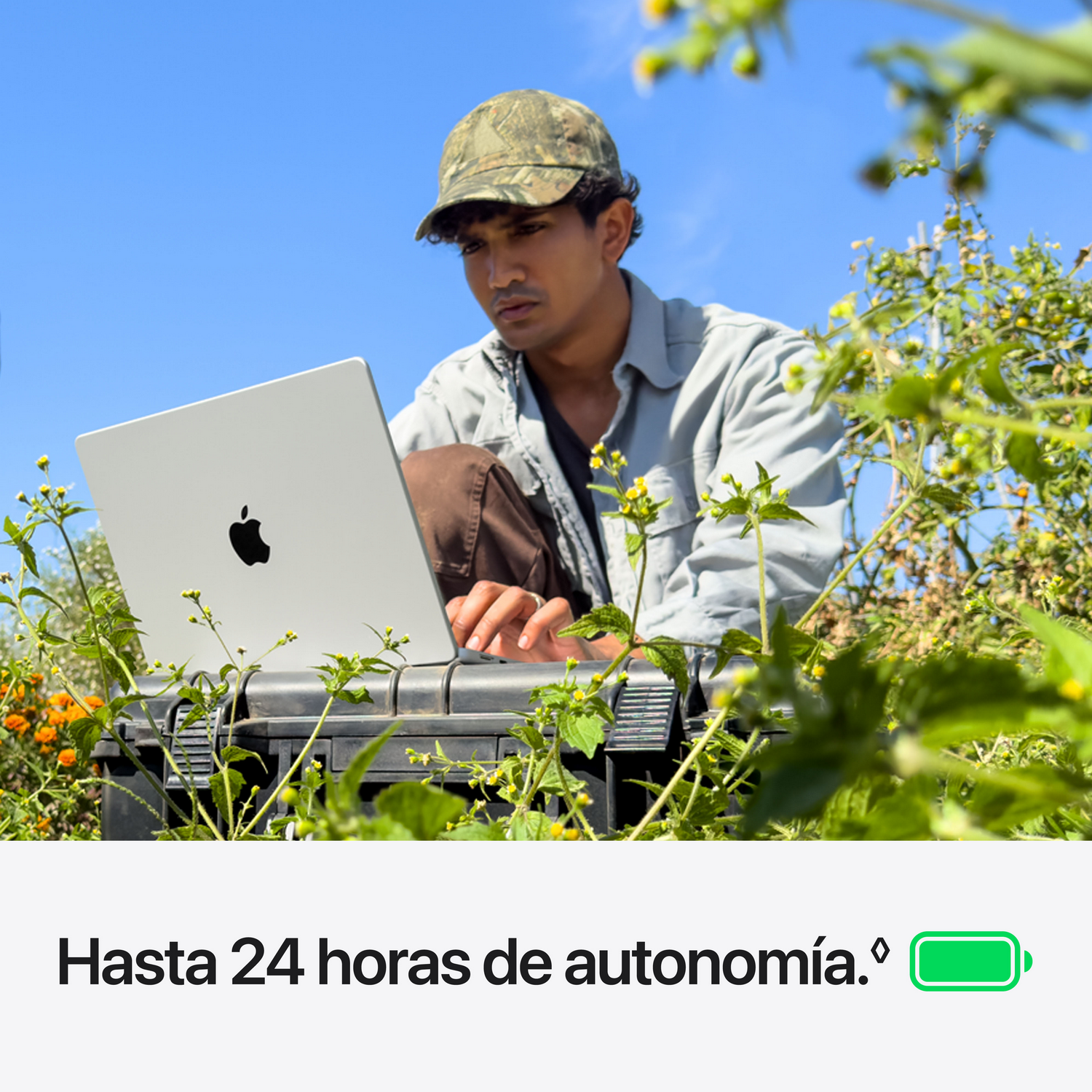 Un hombre en un campo usando una laptop sobre una caja negra, bajo un cielo azul, con texto e icono de batería abajo.