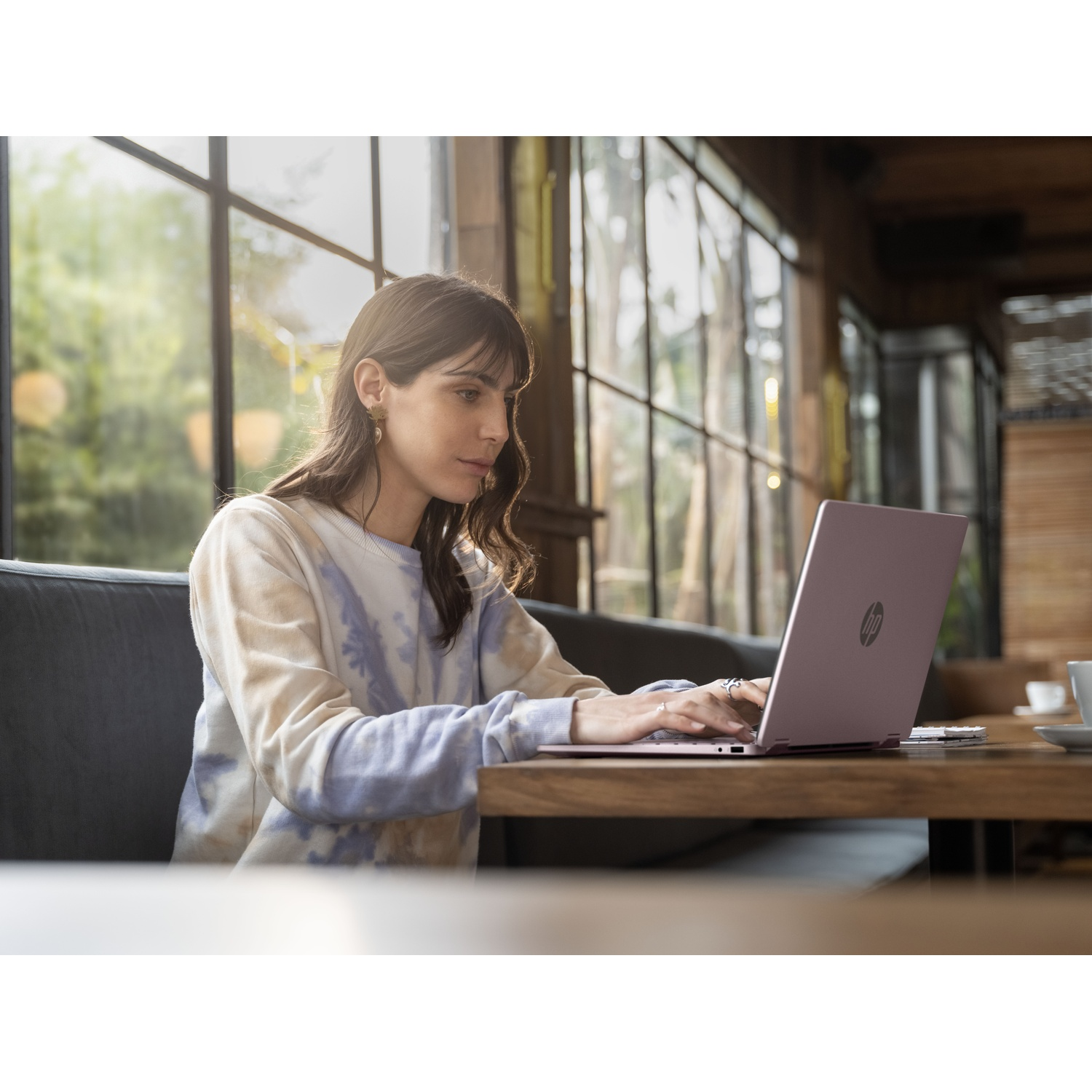 Een vrouw met lang donker haar zit aan een houten tafel in een café te typen op een laptop.