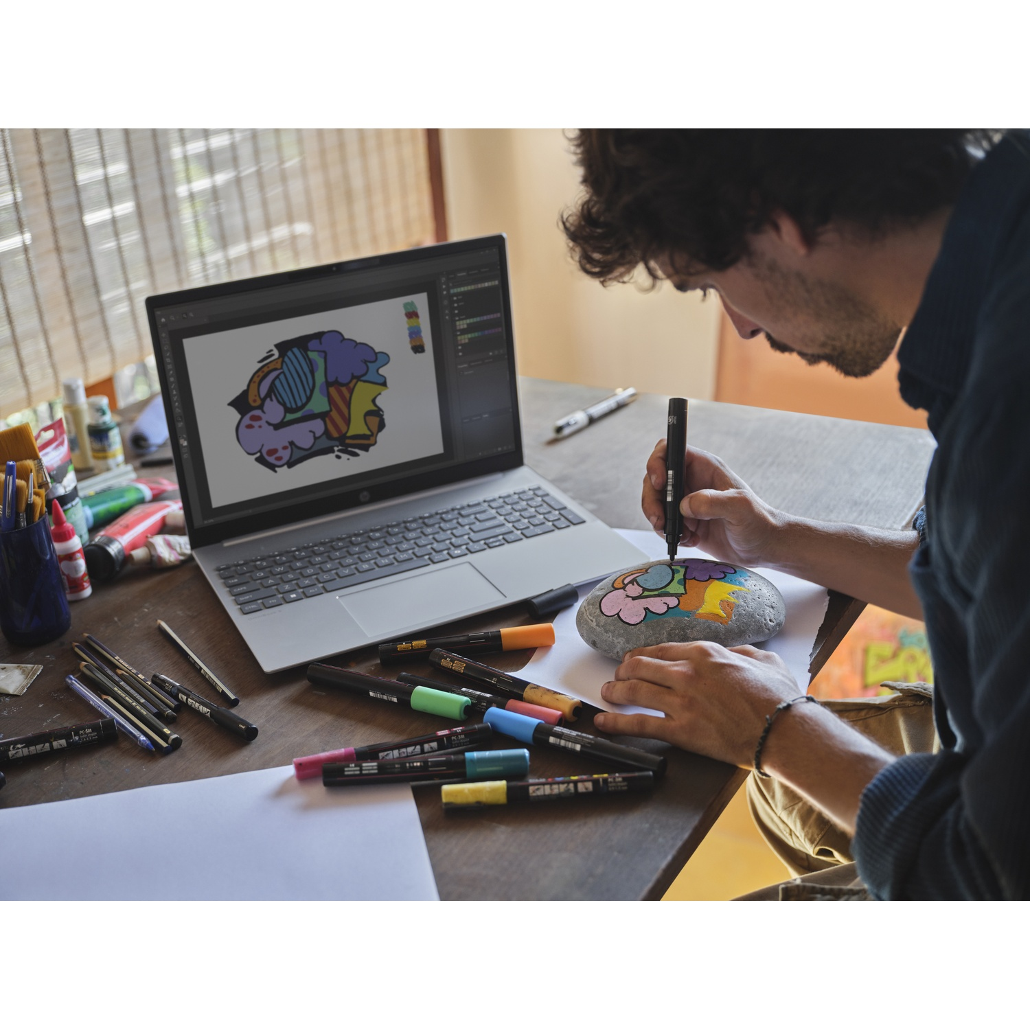 Een man aan een houten tafel gebruikt een marker om een kleurrijk ontwerp op een steen te tekenen voor een laptop.